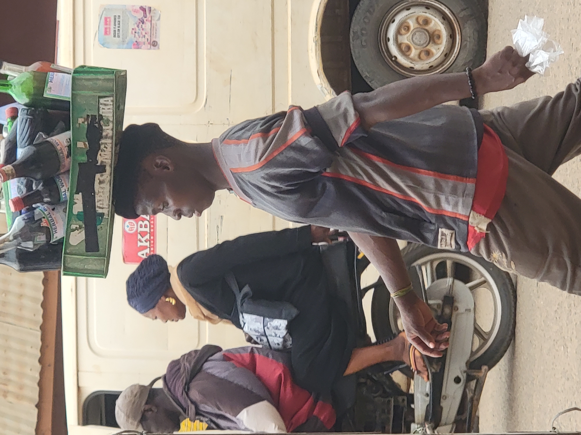 The photo was taken outdoors in Maiburiji, Nigeria. A Black man stands to the right of the frame, holding a balled-up piece of white paper. On his head, a green crate stacked with bottles of drinks is being carried on his head. Another man is seated on a motorcycle, wearing black and a navy blue hat. Behind the man on the motorcycle sits another man with a gray cap and a maroon top. Behind the men is a cream-colored vehicle.