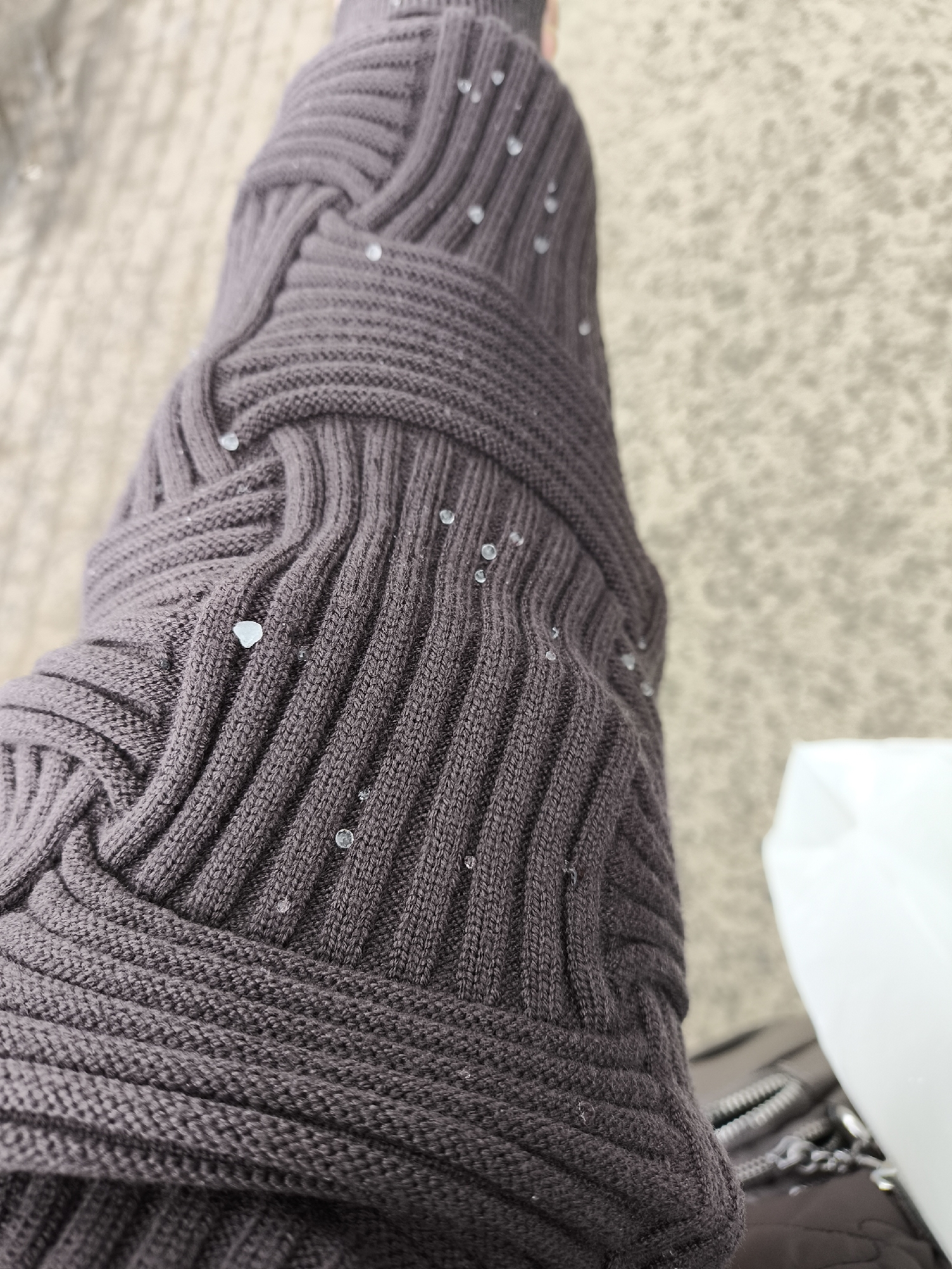 A close-up shot depicts a sleeve of a dark grey knitted sweater adorned with small, clear, bead-like embellishments scattered across its surface. The textured fabric features a ribbed pattern, with the knitting style presenting a series of intertwining loops and strands. The background is blurred and out of focus, showing muted tones. The image also captures a portion of a white object and part of what appears to be a handbag with a zipper and chain detail in the lower right corner. The setting or location of the image, Khimki, Russia, is not visually apparent within the photograph itself.