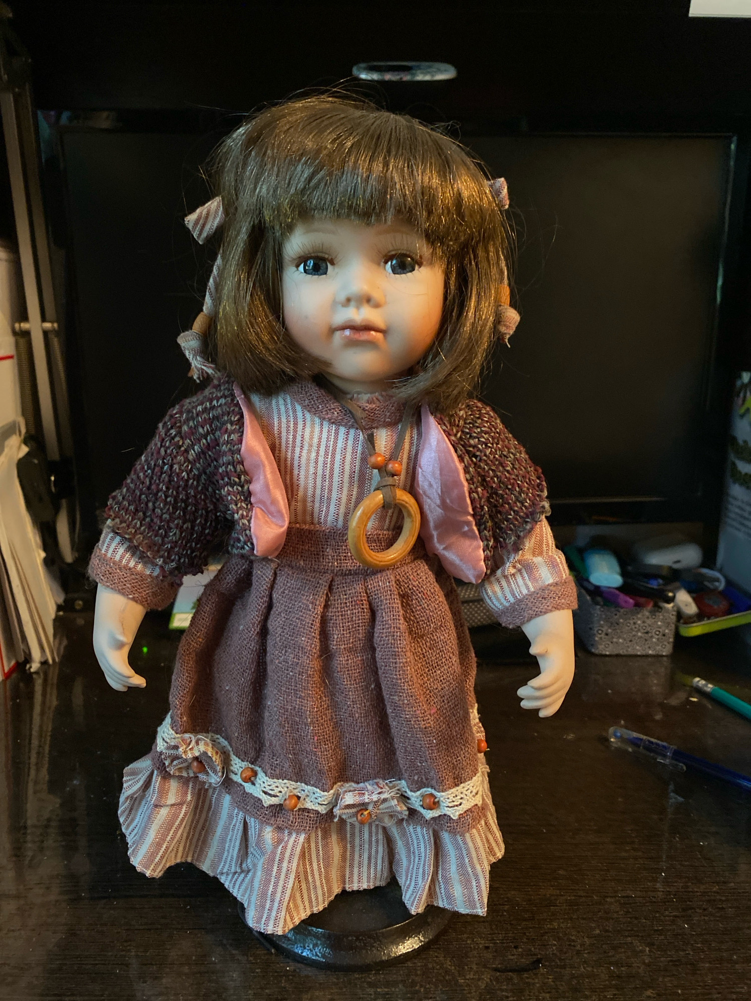 This is a full-body, slightly low-angle, medium shot of a porcelain doll standing on a dark wooden surface. The doll is positioned centrally and facing forward. It has light brown hair styled with bangs and two pigtails tied with small striped bows. The doll has blue eyes, rosy cheeks, and a neutral expression. It is wearing a layered outfit consisting of a striped underdress with a ruffled hem, a darker textured fabric overdress with a ruffled hem trimmed with white lace and small orange beads, and a knitted dark maroon shrug. A pink satin ribbon is visible peeking out from the neckline. A wooden ring necklace with two wooden beads hangs around the doll's neck.
The background is dark and out of focus, suggesting a dimly lit indoor environment. Behind the doll, a dark electronic screen is visible, possibly a monitor or television. To the left of the doll, a stack of papers or books is leaning against something. To the right, a small grey container filled with various items, including pens, markers, and other small objects, is on the surface. A blue pen and a green pencil are also visible on the wooden surface in the foreground. The lighting is focused on the doll, with shadows cast on the surface and the background.
The image appears to be taken in a domestic setting, likely a desk or shelf. The specific location in Ivanteyevka, Russia, cannot be determined from the visual information present in the image.