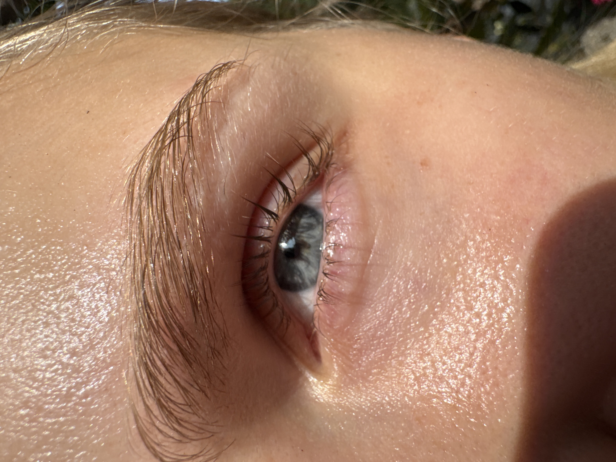 This is a close-up, eye-level shot of a person's eye and eyebrow. The skin appears to be lightly sweating or dewy, with sunlight reflecting off of it. The eyebrow is light brown and well-defined. The eye is blue with dark eyelashes. The background is blurred and out of focus, suggesting an outdoor environment with some foliage. There are no identifiable landmarks or details that would place this image specifically in Nam Nha Trang Ward, Vietnam.