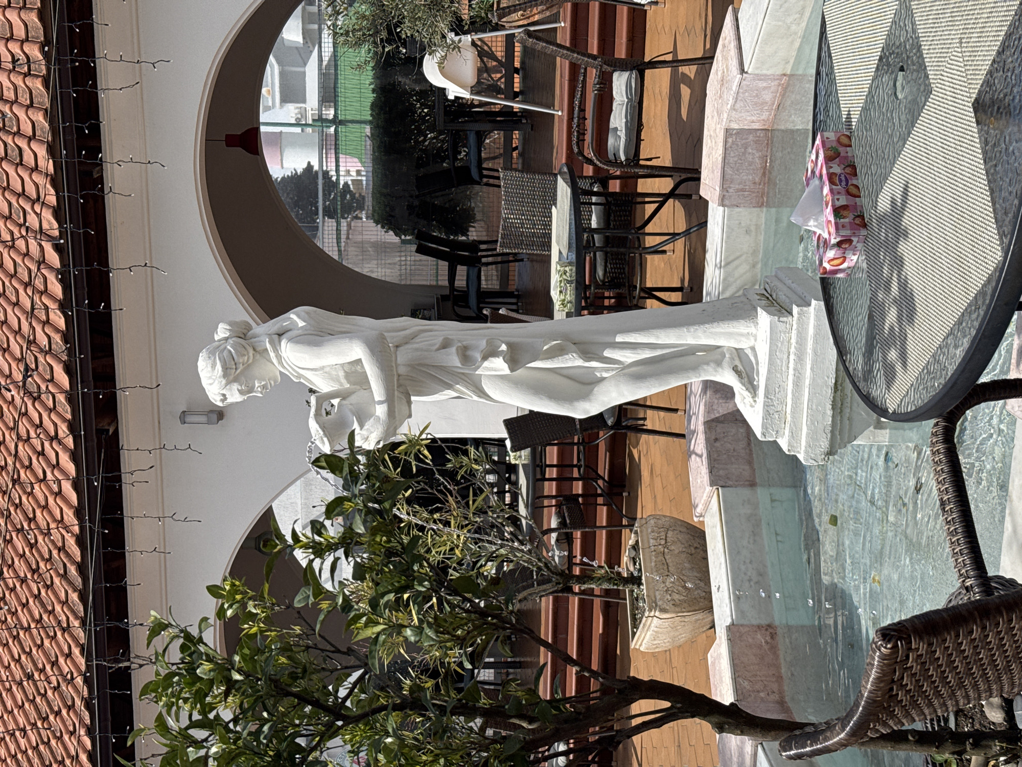 Here is a description of the image:
The image depicts an outdoor scene, possibly a patio or garden setting. A white marble statue of a draped female figure stands prominently in the foreground. To the right of the statue is a glass-topped table with a box of tissues on it, and a wicker chair. Behind the statue, a series of arches frame a view of additional outdoor seating, with tables and chairs arranged on a brick-paved area. A potted tree with green leaves is positioned in the lower left corner, partially obscuring the statue. The setting is outdoors during daylight, and there's a building with a red tile roof visible in the background with decorative lights. The location may be in the city of Sochi, Russia. The statue sits atop a tiered base, adding to its prominence in the scene.