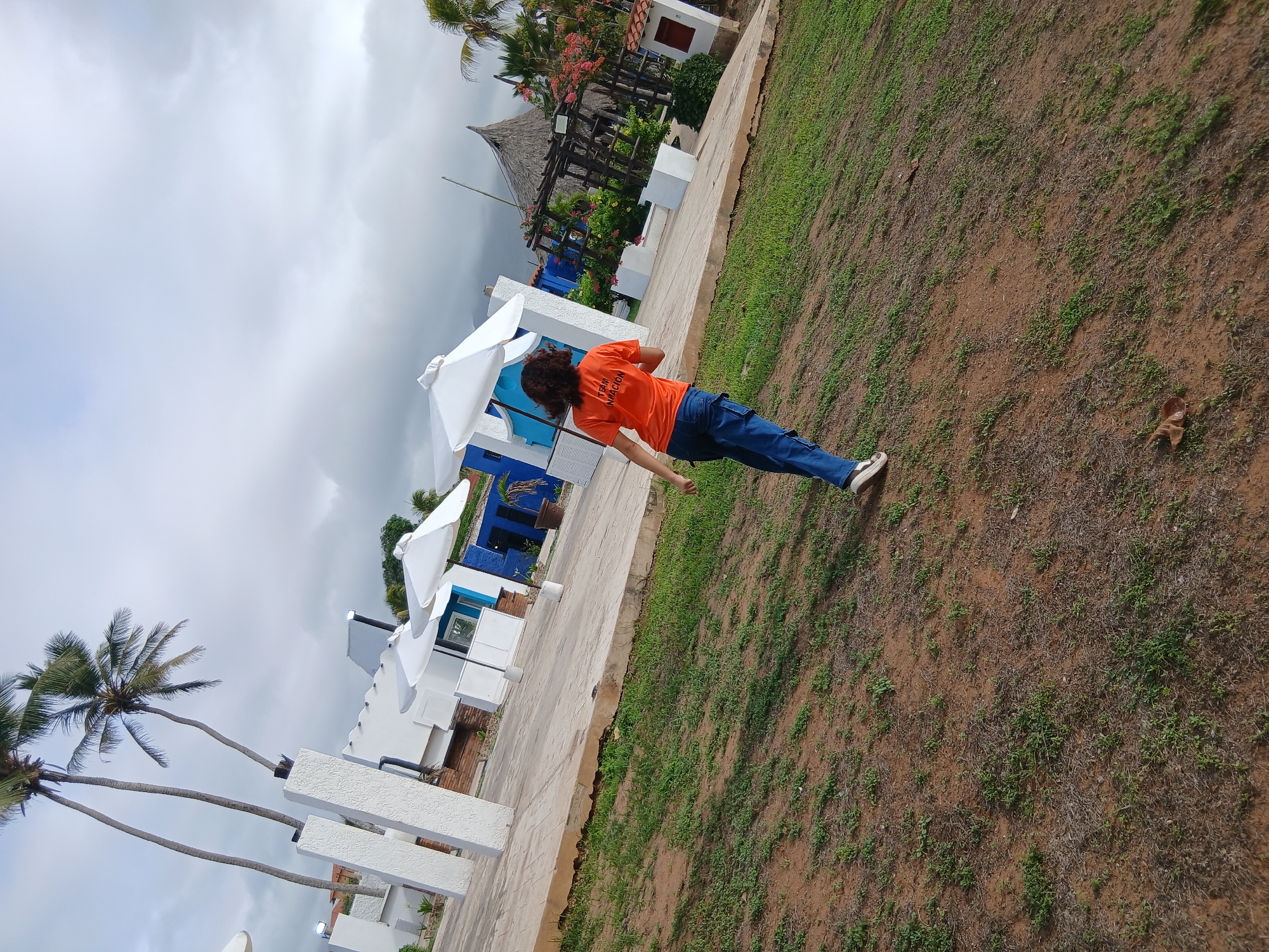 The image depicts an outdoor scene on what appears to be a cloudy day. A young person, seen from behind, is walking away from the camera on a grassy area. They are wearing an orange t-shirt with text on the back that reads "TEAM INFLACION," and blue pants with sneakers. The background shows a paved area with several white structures, some with blue accents, and white umbrellas. Tall palm trees are visible to the left. To the right, there is a thatched-roof structure and more vegetation. The overall environment suggests a resort or recreational area. The location is identified as Playa El Agua, Venezuela.