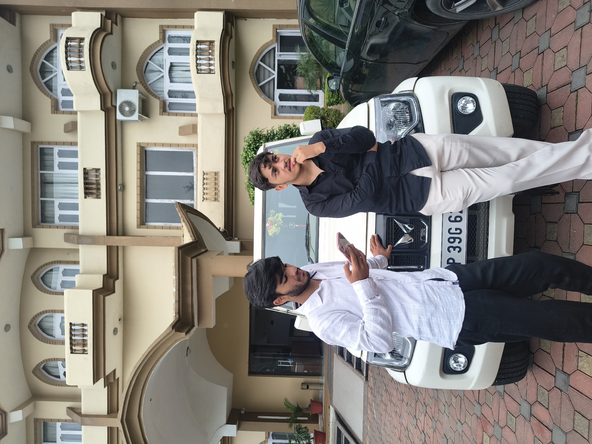 In Khaniara, India, two young men stand in front of a cream-colored building. A white SUV is parked directly in front of them. The man on the left, wearing a white shirt and black pants, is looking down at his phone. The man on the right, in a black shirt and light-colored pants, has his hand on his chin, looking to the right. The building behind them has multiple windows and balconies, and an air conditioning unit is visible on the exterior wall. The ground is paved with interlocking bricks. A black car is partially visible to the right of the white SUV.