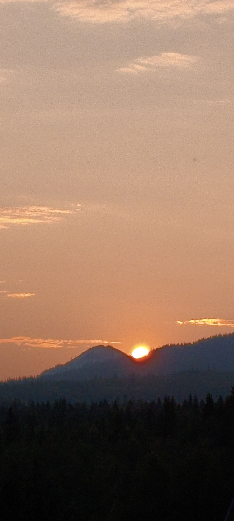 The image shows a sunset over a mountainous landscape. The sun is a bright orb low on the horizon, partially obscured by a dark, silhouetted mountain range. The sky is a gradient of soft orange and peach hues, with a few wispy clouds scattered across the upper portion of the frame. In the foreground, a dense forest of dark trees stretches across the bottom of the image, also silhouetted against the lighter sky. The overall impression is one of natural beauty and tranquility. The location is specified as Zyuratkul', Russia.