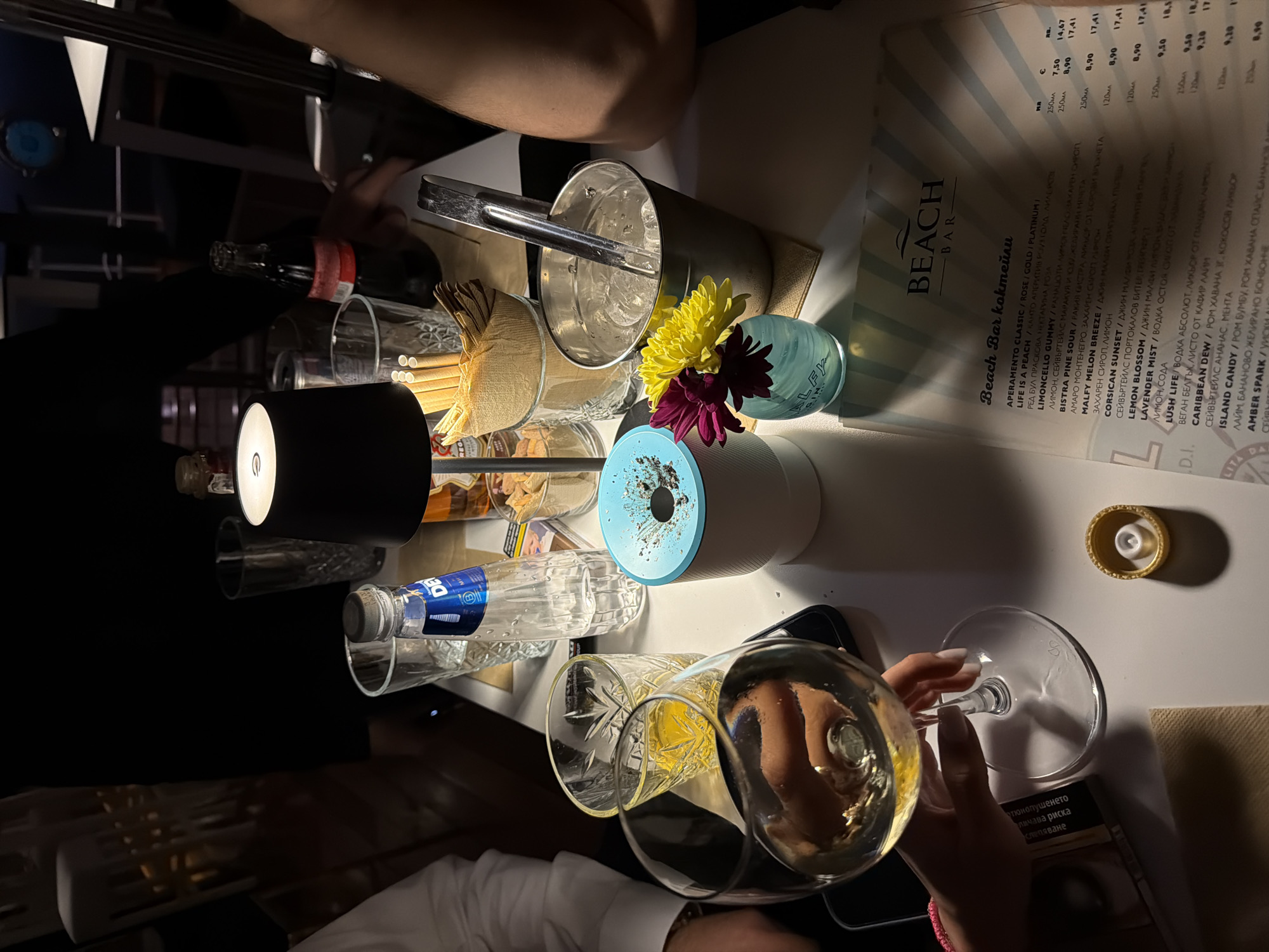 The image depicts a table at a bar, likely in Sofia, Bulgaria, due to the Bulgarian text on a cigarette pack and the drink menu. The "Beach Bar" menu lists cocktails with prices in both an unspecified currency and euros (€). A person is holding a wine glass filled with a light-colored beverage. An ice bucket with ice tongs and flowers next to a clear jar, and a light blue vase is also on the table. Other objects present include a black lamp, several bottles of water and other unidentified drinks, and straws. There are also hands of two people in the image and portions of their arms, along with a white sleeve and a pink bracelet.