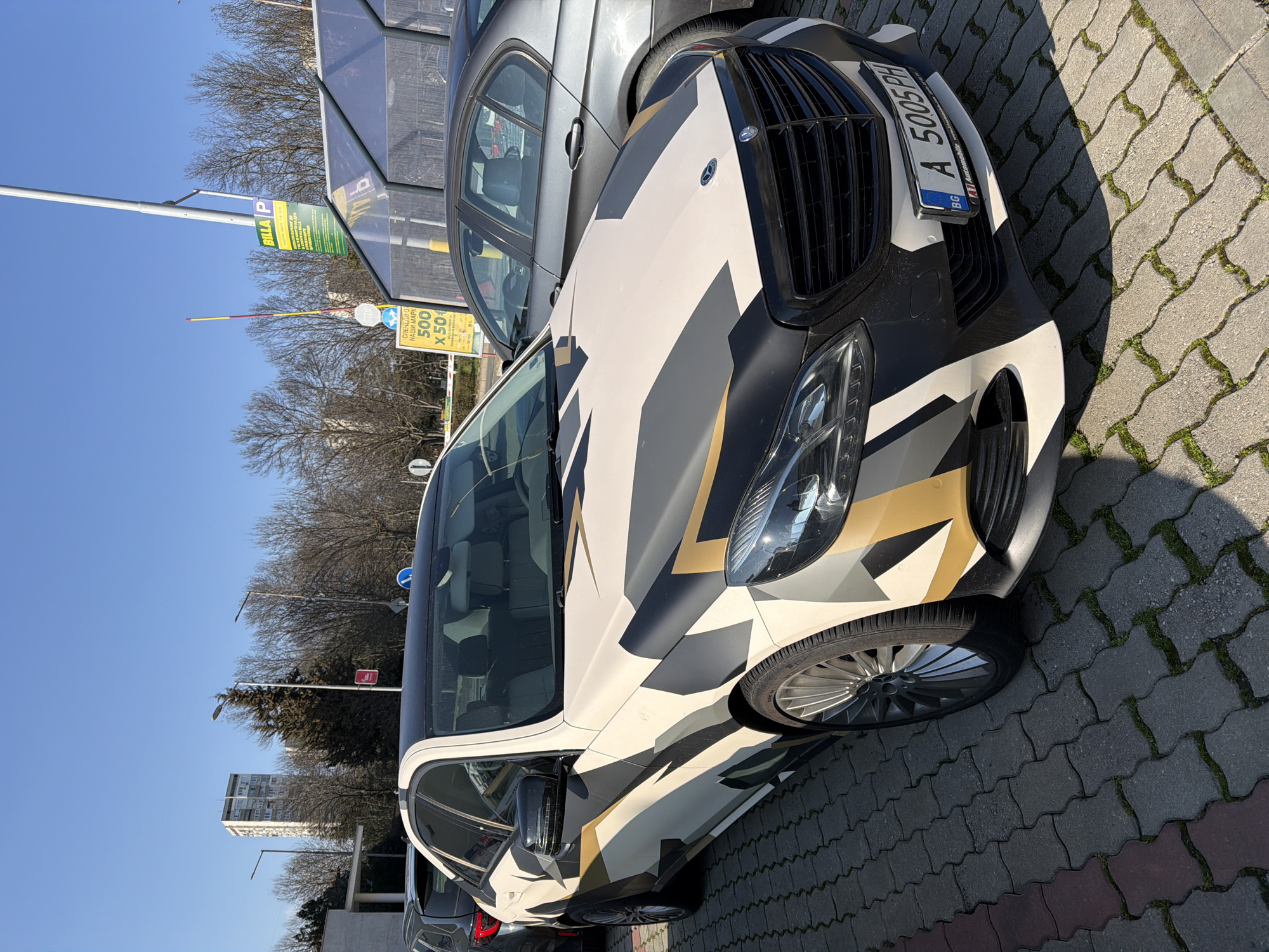 An overhead-angle shot captures a custom-wrapped Mercedes-Benz E-Class sedan parked on interlocking grey concrete pavers in what appears to be a supermarket parking lot. The vehicle, a W212 facelift model, features a striking geometric camouflage wrap in off-white, dark grey, and gold/tan, with a contrasting black roof. Its front license plate, "BG A 5005 PH," identifies it as registered in Burgas, Bulgaria. The car is equipped with multi-spoke alloy wheels.
To the immediate right, a dark grey sedan (possibly a BMW E90 series) is partially visible. To the left, part of a darker SUV or hatchback is present. The parking surface exhibits some moss or grass growth between the pavers.
The background features bright daylight under a clear blue sky, casting distinct shadows. A tall pole displays a prominent yellow "BILLA P" sign, indicating a Billa supermarket and parking. Below it, another yellow sign advertises a promotion involving "500 X 50лв" (500 x 50 BGN). A red-and-yellow striped parking barrier arm is partially visible. Road signs, including a blue circular mandatory direction sign and a red circular warning/prohibitory sign, are also present. The scene includes bare deciduous trees, an evergreen conifer, streetlights, and a distant, multi-story residential or office building. A transparent or translucent canopy structure is visible to the right of the parked vehicles. The location is a Billa supermarket parking area within Burgas, Bulgaria.