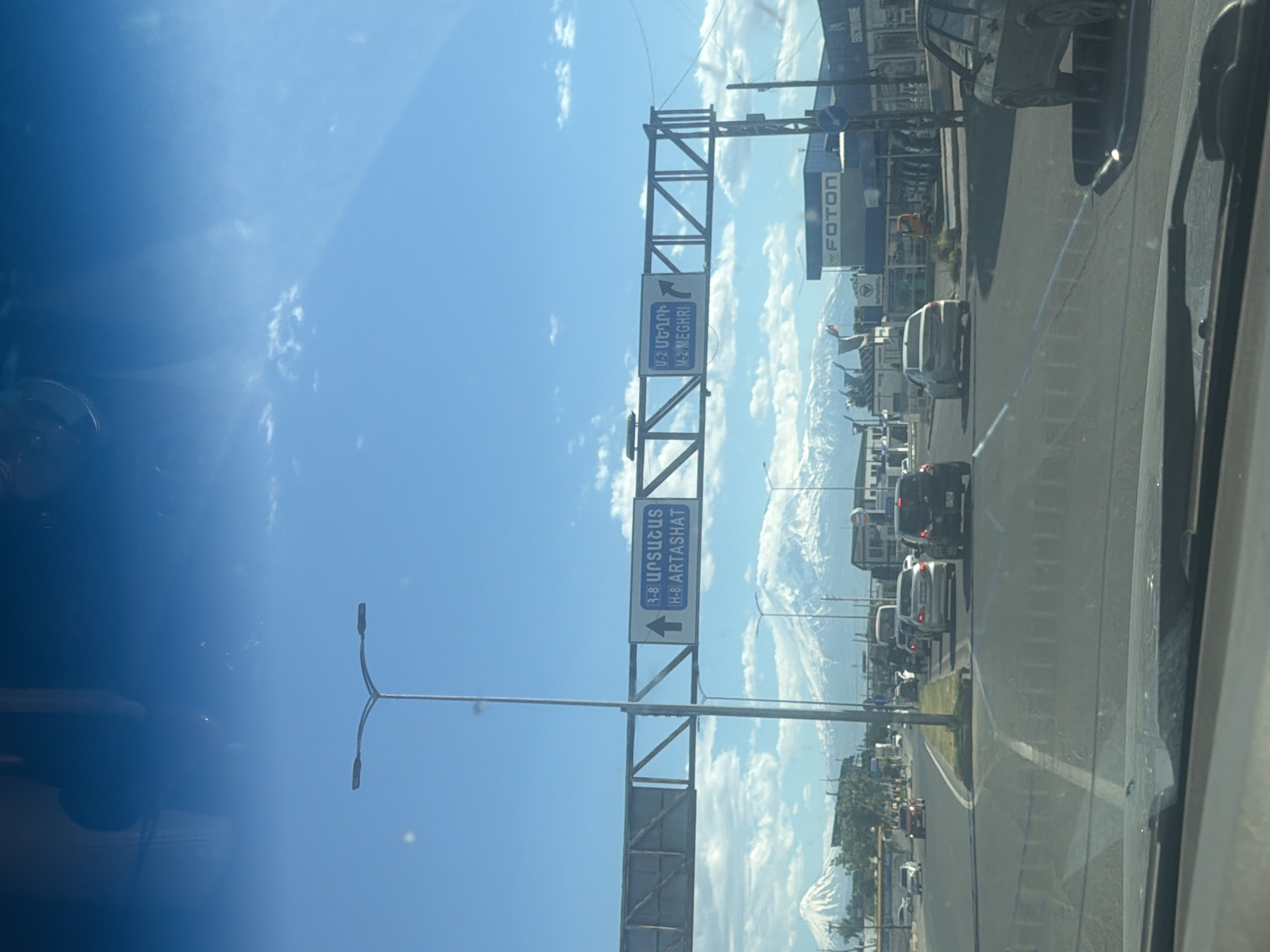 Captured from within a vehicle, the image depicts a road scene under a partially cloudy, bright sky. The road, with visible lane markings, is busy with vehicular traffic. Above the road, a metal gantry displays road signs in Armenian script and English. The signs indicate directions to H-8 Artashat and H-2 Meghri. In the backdrop, a mountainous landscape merges with the cityscape. The environment is consistent with a highway intersection in an urban area. The location is somewhere in Armenia.