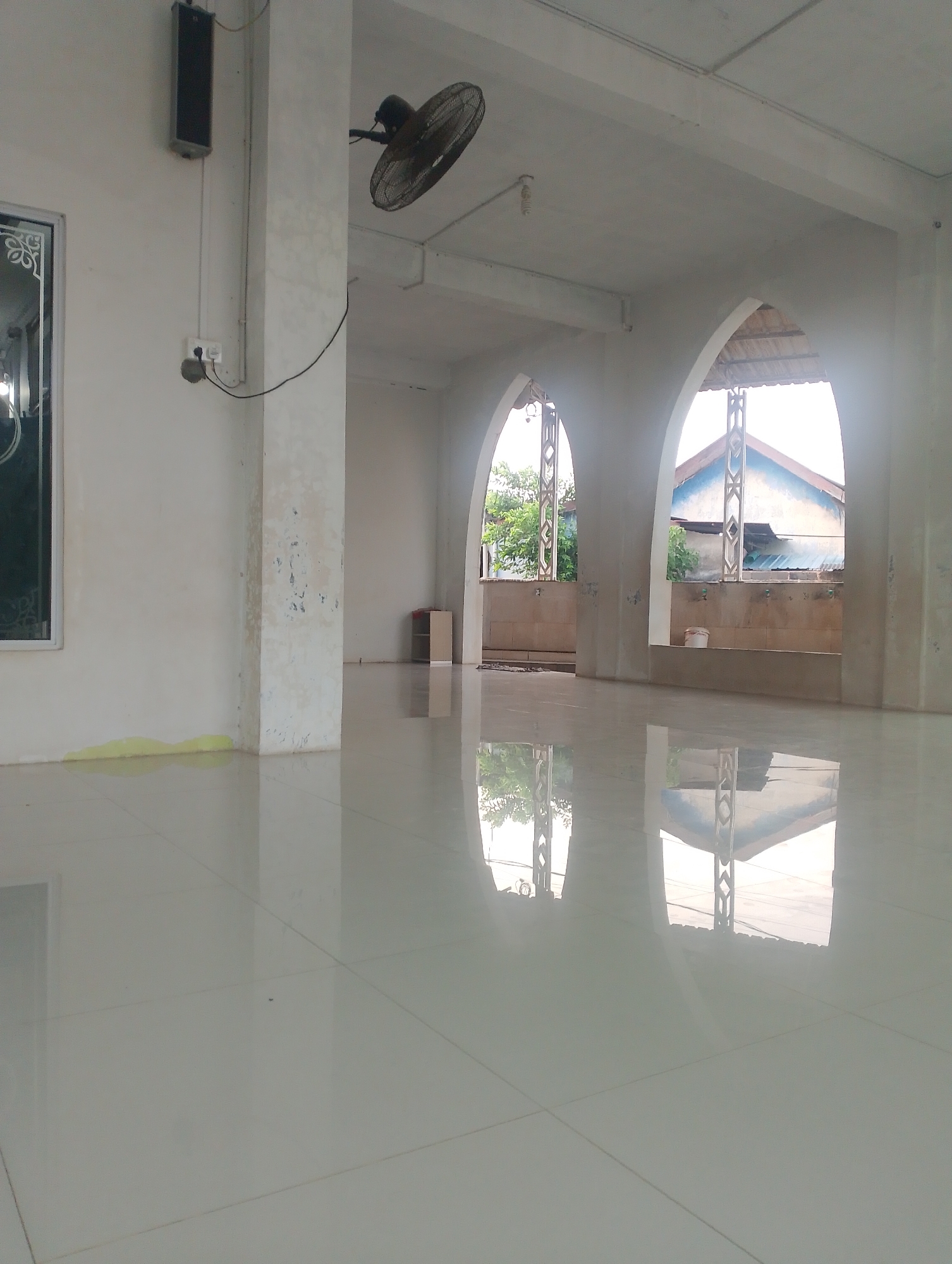 The image depicts the interior of an unfinished or sparsely furnished building, possibly a prayer hall or community space, in Batam, Indonesia. The room has a high ceiling and is illuminated by natural light filtering through two large arched openings. The walls are painted white, with some signs of wear and tear. A black ceiling fan is visible in the upper left of the frame. A speaker is mounted on the wall to the left of the fan, with electrical wiring running down to an outlet. To the left, a partially visible window with decorative framing is present.
The floor is made of large, glossy white tiles that create strong reflections of the room and the external environment. In the foreground, a bright yellow liquid stain is visible on the floor near a pillar.
Through the arched openings, an outdoor scene is visible, showing green trees, residential buildings with blue roofs, and power lines. The architecture of the arches suggests a tropical or Southeast Asian style. One arch reveals a trough with faucets along the wall, possibly for ablution. A white bucket is placed in front of the trough in the right arch.
The overall impression is one of a functional space under construction or preparation, with basic amenities and a focus on clean, reflective surfaces. The presence of a prayer hall-like setting and the location in Batam, Indonesia, contribute to the contextual understanding of the scene. There are no people visible in the image.