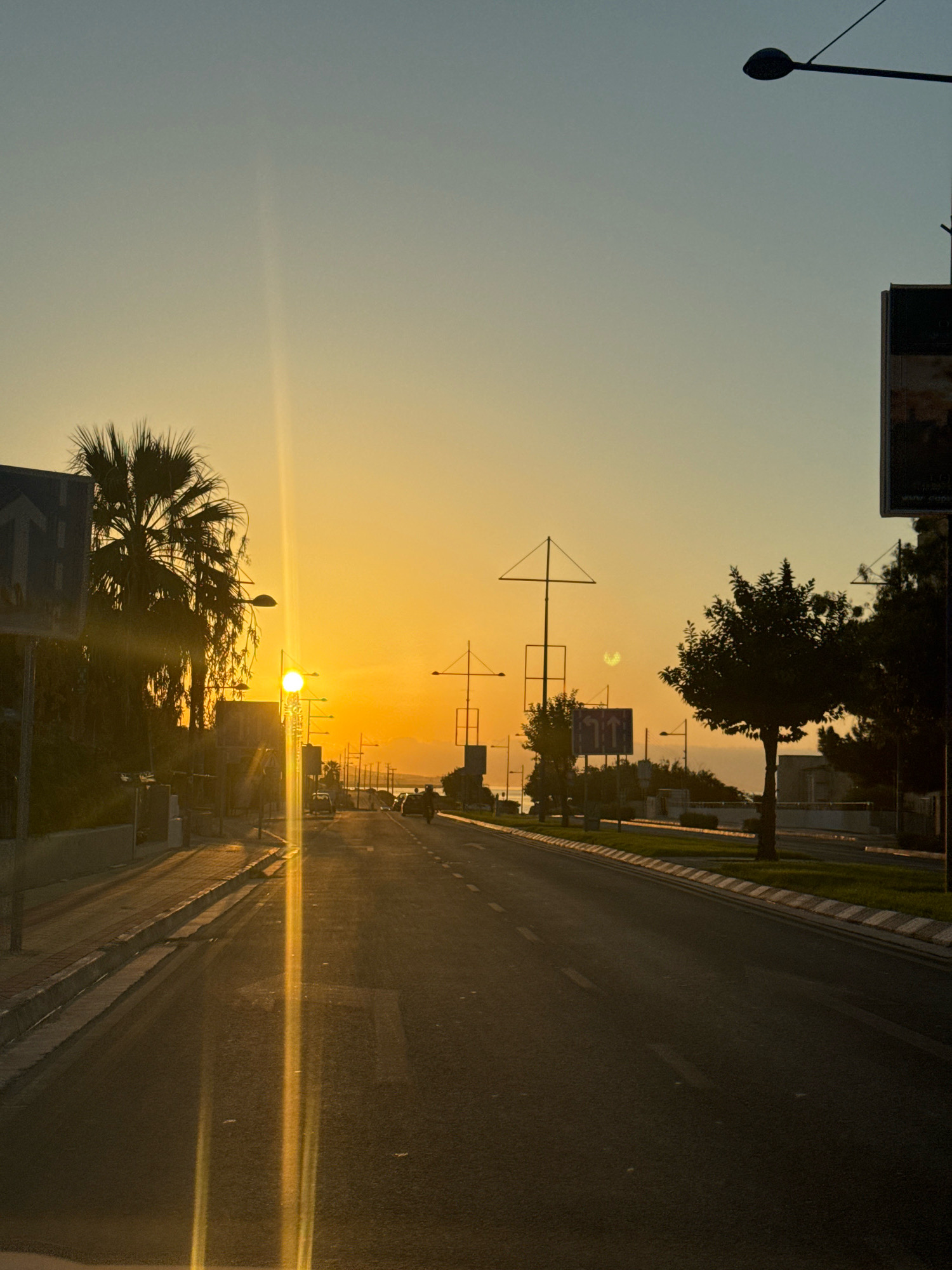 The image depicts a street in Agios Tychona Community, Cyprus, at sunset. The sky transitions from a pale blue at the top to a vibrant orange and yellow towards the horizon, indicating the setting sun. A strong lens flare from the sun creates vertical streaks of light across the image, predominantly on the left side.
On the left side of the street, a sidewalk is visible, bordered by a low wall. Several palm trees line the sidewalk, their fronds silhouetted against the bright sky. A blue traffic sign with white arrows is also present. Beyond the sidewalk, buildings and more palm trees can be seen.
The road itself is asphalt, with white lane markings. Several vehicles, including cars and a motorcycle, are driving on the road, heading away from the viewer. Streetlights are positioned along the road.
In the background, there appears to be a body of water, likely the Mediterranean Sea, with distant hills or landforms on the horizon. The overall scene is one of a coastal town at dusk, with the warm glow of the sunset illuminating the environment.