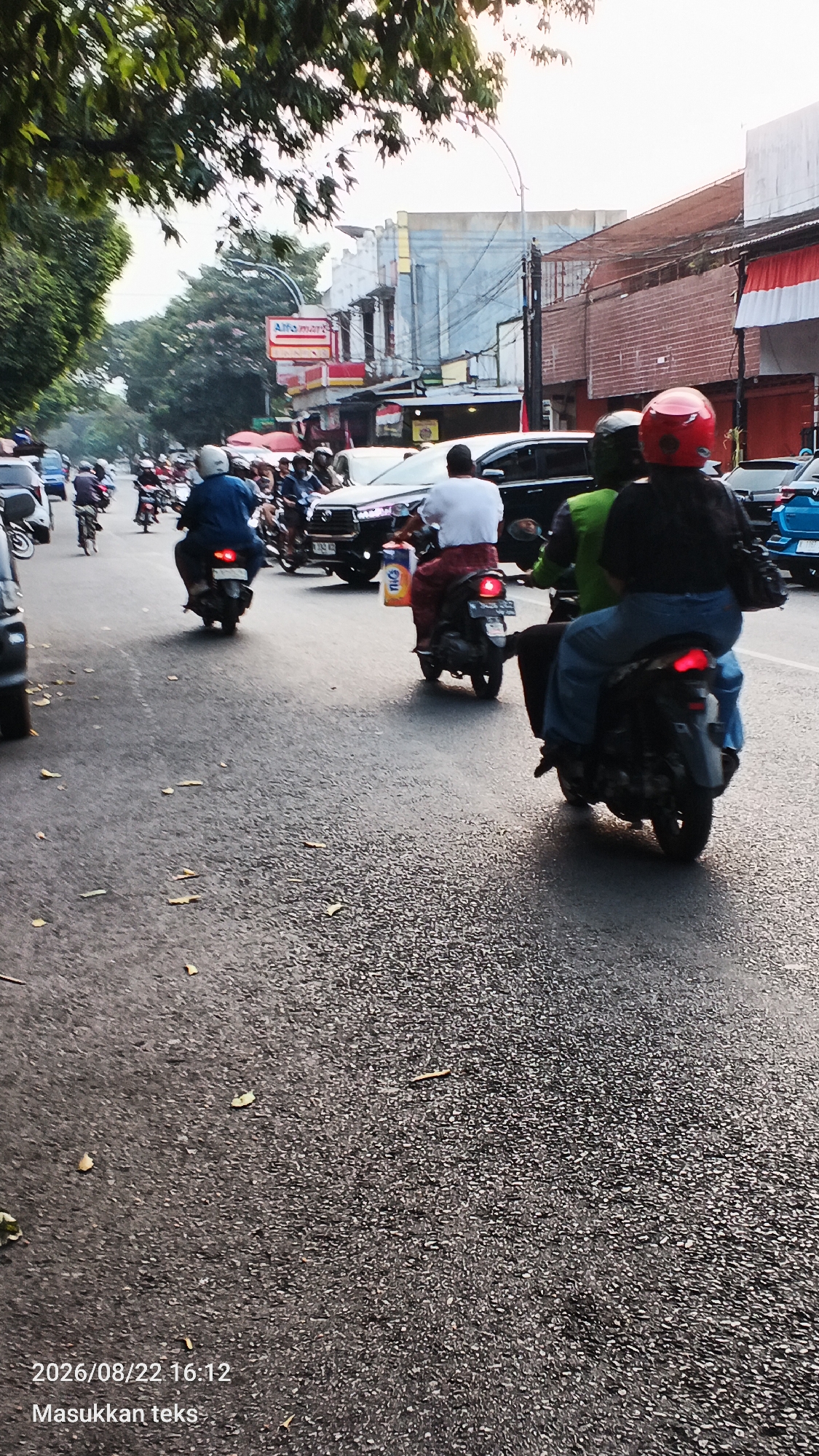 The image depicts a street scene in Malang, Indonesia, likely during the late afternoon, as indicated by the sunlight casting shadows and the timestamp "2026/08/22 16:12". The road is asphalt and appears wet or recently rained on, with scattered leaves on the surface.
The foreground shows the pavement with a timestamp and the text "Masukkan teks" (which translates to "Enter text" in Indonesian) superimposed in the bottom left corner.
In the middle ground, a busy street is visible with numerous motorcycles and a few cars. Several individuals are riding motorcycles, some with passengers. The riders are mostly wearing helmets, with one rider wearing a red helmet. One motorcycle in the center carries a person in a white shirt and another person in a green jacket. Another motorcycle further back has a rider in a blue shirt. A black SUV is also present in the traffic.
The background features buildings lining the street, including a building with a prominent red and white sign that appears to be an "Alfamart" convenience store. Other buildings have a mix of architectural styles, some with balconies and awnings, and there are power lines and street lights visible. Trees with dense foliage overhang the street from the left side of the frame, casting shadows on the road. The overall impression is of a typical urban street scene in Indonesia with moderate traffic.