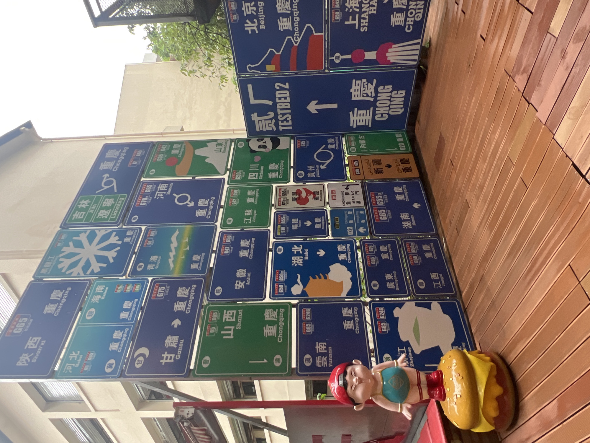 A street art installation featuring numerous road signs pointing to various Chinese provinces and cities, with "Chongqing" appearing frequently, is set up on a wooden deck. In the foreground, a small, chubby figurine wearing a red cap and a turquoise outfit stands on a large, stylized hamburger. The backdrop consists of a building with white and beige siding and windows. The signs are a mix of blue and green, with Chinese characters and some English translations. Some signs have highway numbers like G65 and G75. One sign prominently reads "TESTBED 2" with an upward arrow. The overall scene evokes a whimsical and culturally rich environment.