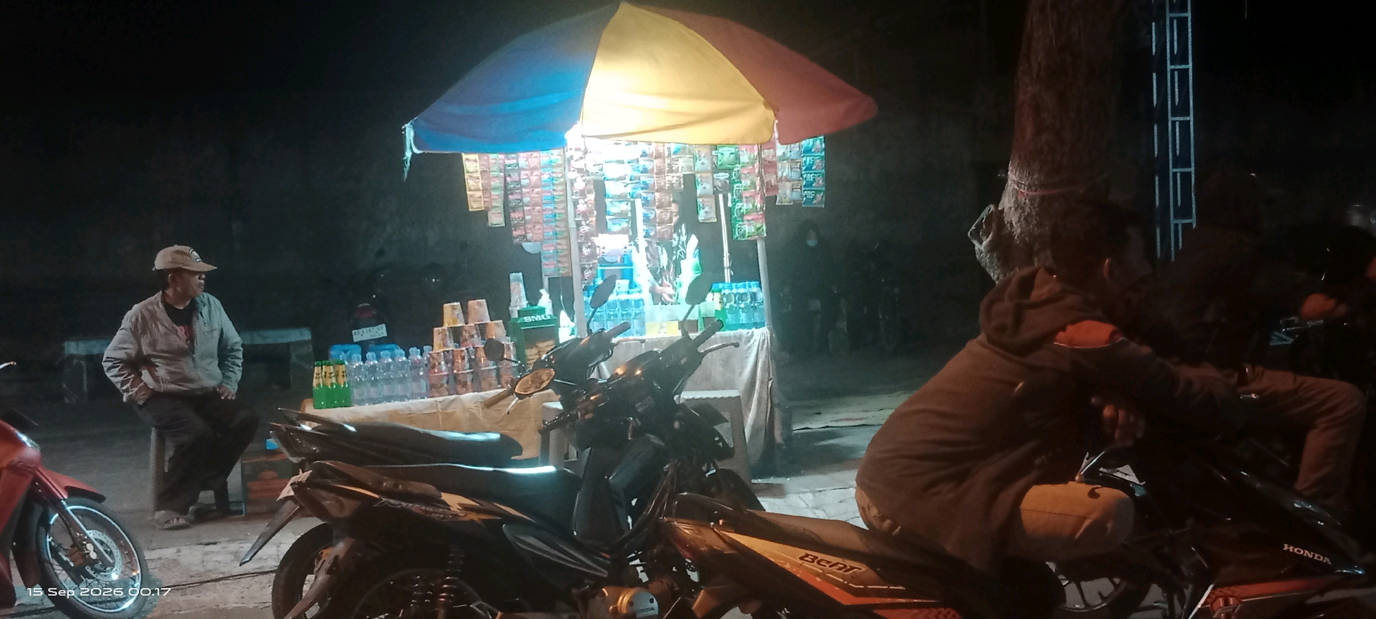 The image depicts a night scene in Klaten, Indonesia, featuring a roadside stall selling drinks and snacks. A man wearing a light-colored jacket and a cap is seated on a stool. In the foreground, two motorcycles are parked. The stall is covered by a colorful umbrella and displays rows of packaged goods. Several bottles of drinks are arranged on a table, alongside stacks of paper cups. The environment suggests a public area, possibly a street or a market, with dimly lit surroundings and a large tree trunk visible to the right. A second individual is seated on another motorcycle in the right portion of the frame. The timestamp at the bottom left indicates September 15, 2026, at 00:17.