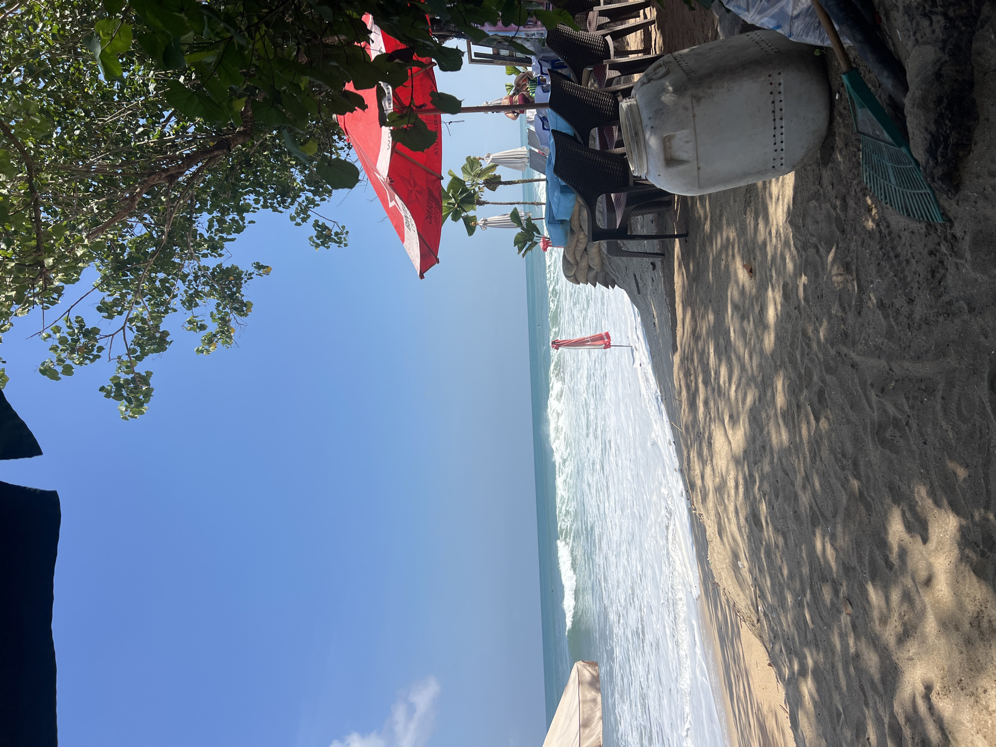 This image captures a section of the Kuta beach in Indonesia, a popular tourist destination known for its vibrant atmosphere. The scene depicts a beachside seating area, likely part of a restaurant or cafe, with tables and chairs set up under a large red umbrella. In the background, the turquoise waters of the ocean meet the sandy shore, with gentle waves rolling in. Palm trees and lush greenery add to the tropical ambiance. A large white plastic container and a rake are visible in the foreground, suggesting preparations for or cleanup after a busy day. A few people are present, seated at the tables or walking along the beach, enjoying the sunny weather and scenic views. The overall impression is one of a relaxed and lively beach scene, characteristic of Kuta's popular tourist spots.