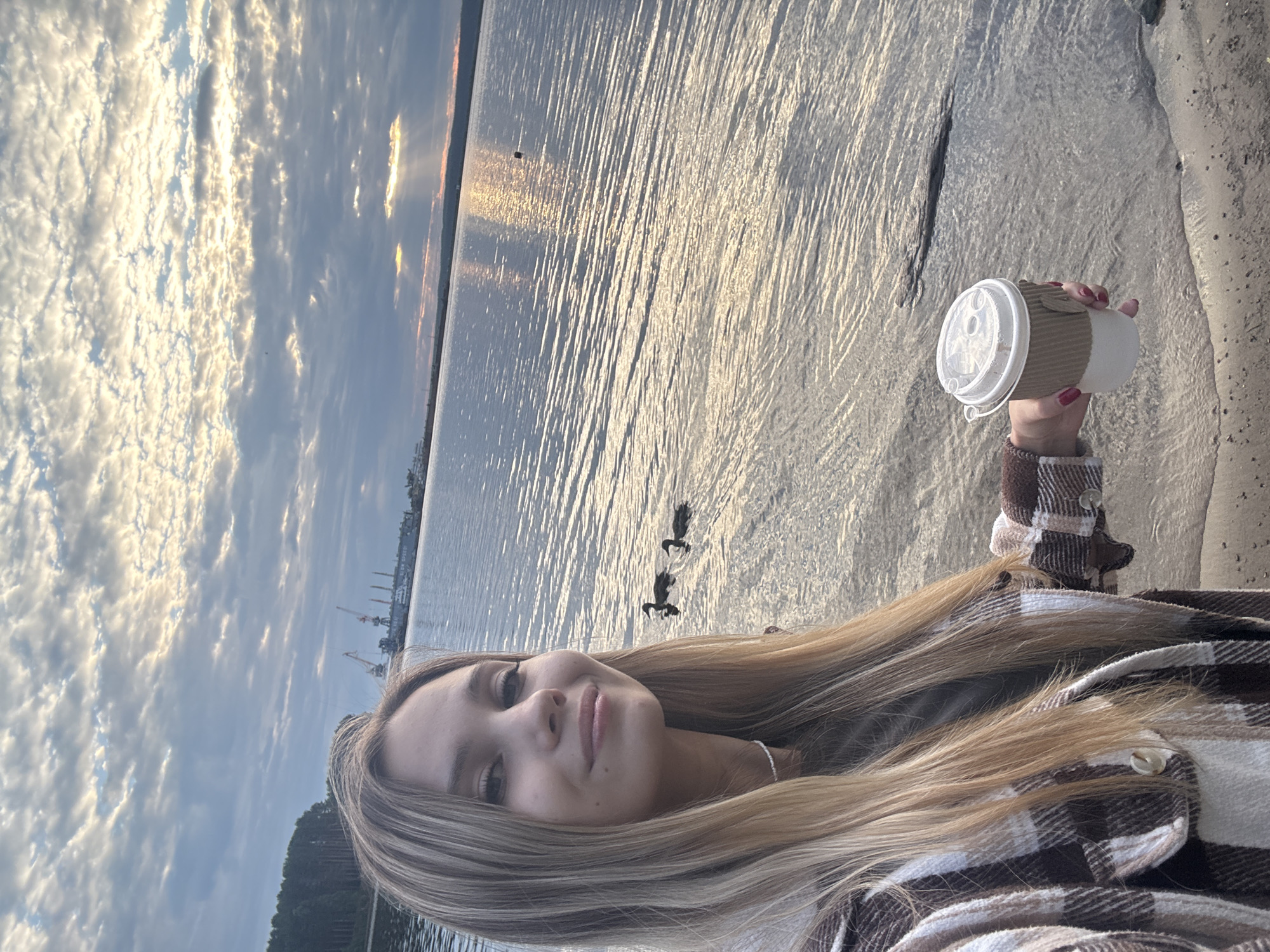 A young woman with long blonde hair takes a selfie by the water, holding a takeaway coffee cup. She is wearing a brown and white plaid shirt. The sun is setting, casting a golden glow on the water and clouds. Two ducks swim in the distance. In the background, industrial cranes are visible, suggesting a port or industrial area. The overall mood is peaceful and serene. The image is oriented vertically.