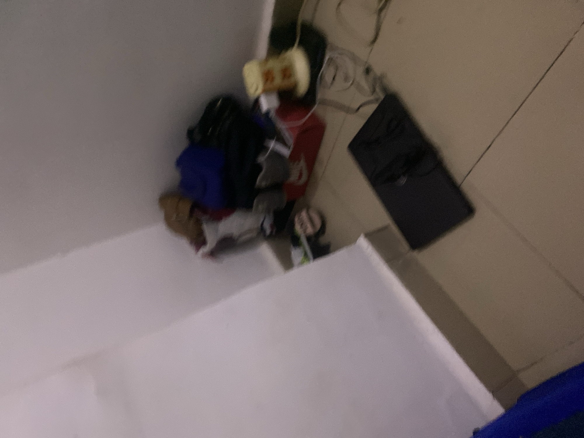 The image shows a dimly lit corner of a room. A white wall meets a tiled floor. In the corner, a pile of items includes a red box, what appear to be shoes, and several bags or folded fabric items in blue and dark colors. To the right, a small lamp with a yellowish-orange patterned shade sits on a black surface, next to a glass and some wires. In the foreground, a black laptop computer is open on the floor, with a mouse positioned on it. The overall scene suggests a disorganized personal space.
