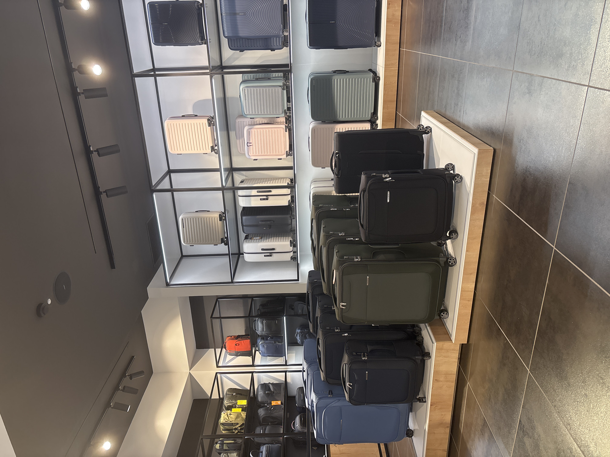 This image captures the interior of a well-lit retail store, showcasing a wide array of luggage. The scene is a modern display area, likely a luggage or travel goods store, situated in Rome, Italy, according to the provided context.
The primary subjects are numerous suitcases and travel bags, arranged on distinct display units. On the left side of the frame (when viewing the original image upright), hard-shell suitcases are presented within minimalist black-framed shelves with white bases. These cases feature various colors, including light pink, white, light blue, mint green, and dark blue, many with textured or ribbed exteriors. Several cases prominently display the "SAMSONITE" brand logo.
On the right, a separate display unit features a collection of soft-shell suitcases and bags. These are predominantly in darker tones such as black, dark blue, and olive green, with some beige and an orange bag visible further down. All visible luggage appears to be designed for travel, equipped with wheels and handles.
The store's interior features a dark grey ceiling with multiple bright track lights illuminating the merchandise. The floor is covered in large, dark grey or charcoal-colored tiles with lighter grout lines, creating a clean and contemporary aesthetic. No people are visible, and the overall activity depicted is the static display of products for sale. The bright lighting suggests either daytime operation or a well-lit indoor environment. No specific time of day or weather conditions are discernible from the interior shot.