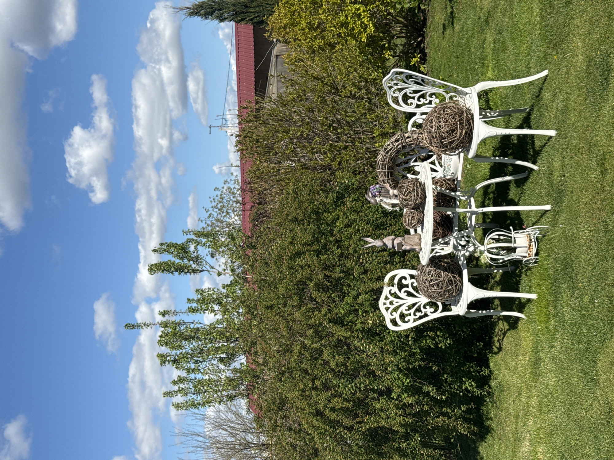 This image captures a serene outdoor garden scene in Pervorechenskoye, Russia, on a bright, sunny day. The focal point is a white, ornate metal garden table surrounded by three matching chairs, all featuring intricate scrollwork and decorative designs.
The table and chairs are adorned with several decorative items, primarily round, woven balls made from natural brown materials, varying in size. One large woven ball rests on a chair to the left, another on the table, and smaller ones are also present. A woven wreath-like decoration is also placed on the table, alongside a small, light-colored decorative figure that appears to be a bird or rabbit. A small white basket or container is visible under the table.
The ground is covered with neatly kept green grass. In the background, a dense wall of lush green bushes and trees creates a natural backdrop. Partially visible behind the foliage is a building with a distinctive reddish-brown corrugated roof. The sky above is a clear, vibrant blue, dotted with scattered, fluffy white cumulus clouds. The bright sunlight casts distinct shadows from the furniture onto the grass, indicating it is daytime. There are no people visible in the image, and no specific activity is taking place, but the arrangement suggests a prepared outdoor seating area for relaxation. No text is visible.