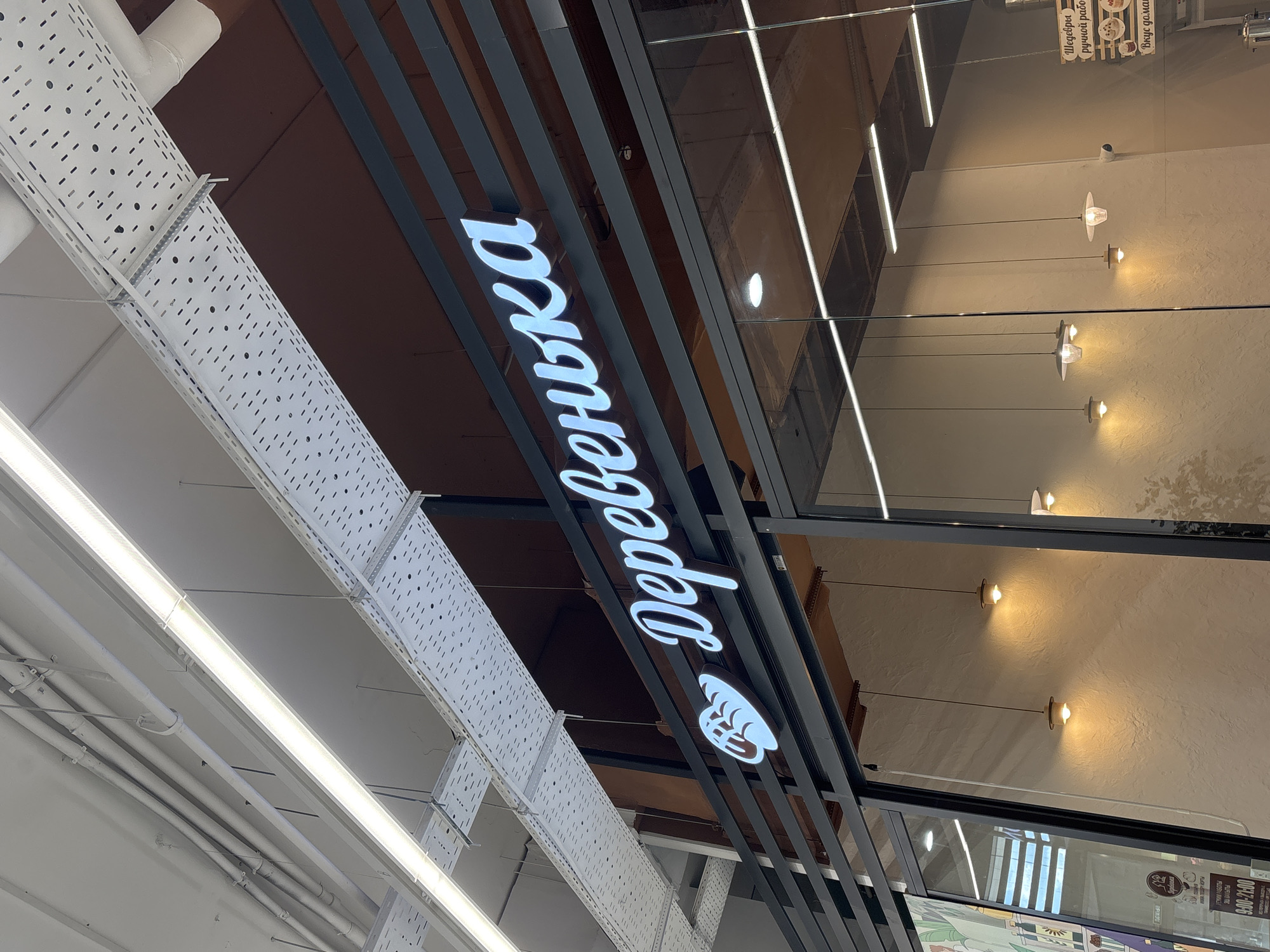 The image shows the exterior of a store with a sign that reads "Деревенька". The sign is illuminated and appears to be made of white lettering with a blue outline, and it features a graphic of a loaf of bread. The store has large glass windows. Inside the store, there are pendant lights hanging from the ceiling. The building is modern, with exposed ceiling structures and industrial-style metal beams supporting the signage. In the lower left corner of the image, there is a poster with text in Russian, including what appears to be operating hours "9:00-21:00". This sign indicates the location is likely a bakery or food establishment. The overall environment suggests an indoor commercial space. Based on the name "Деревенька", which translates to "Village", and typical store branding in Russia, this is likely a chain of bakeries or grocery stores. While the exact location within Naberezhnye Chelny is not provided by the visual elements, the text on the poster and the presence of Russian signage strongly suggest this city in Russia.