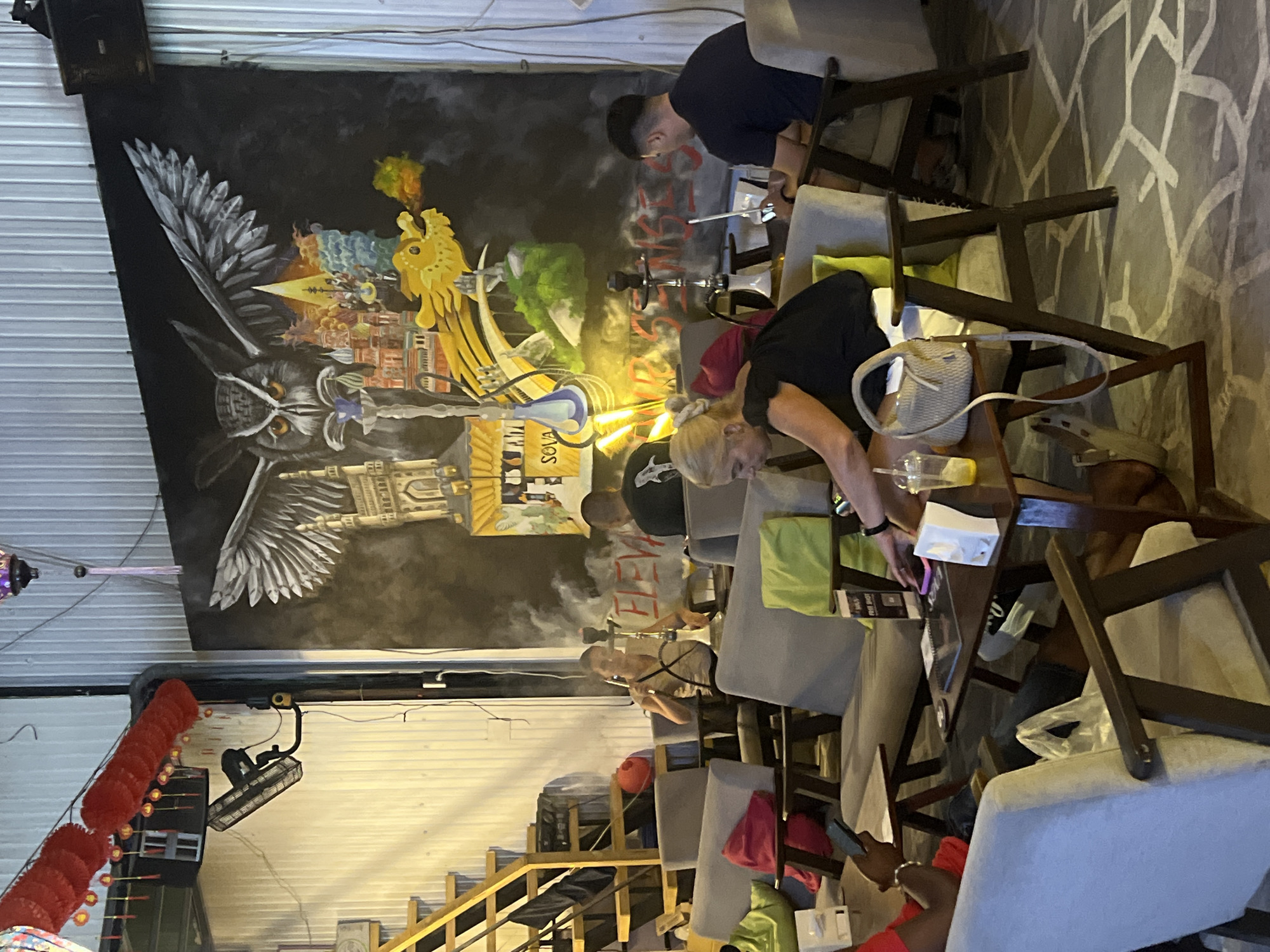The image shows an indoor space in Đà Nẵng, Vietnam, with people seated at tables, apparently in a hookah lounge or similar establishment. The focus is on a group of individuals engaged in smoking hookahs or shisha.
In the background, a large mural covers the wall, depicting an owl with architectural elements integrated into its design. This mural seems to incorporate imagery of cityscapes, dragons, and other symbolic motifs.
There are multiple seating arrangements with patrons smoking. The tables are cluttered with hookah pipes, menus, and personal items like bags and mobile phones. The environment includes decorations such as hanging red lanterns and exposed ductwork or ventilation. A staircase is also visible in the lower portion of the frame. The lighting appears to be a combination of natural light and artificial sources.