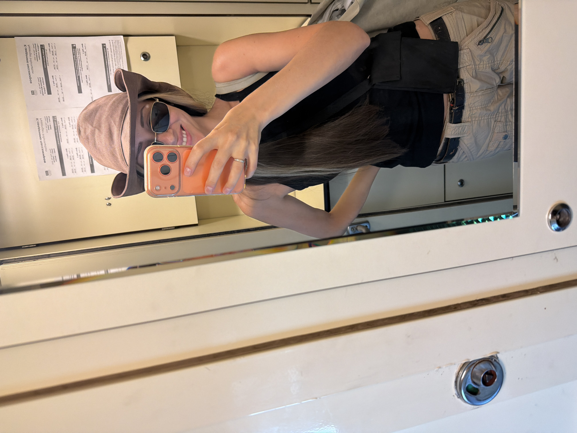 A woman wearing a cowboy hat, sunglasses, a black tank top, and khaki cargo pants takes a selfie in a train car. She is holding an orange smartphone in her right hand and has her left hand on her hip. A black crossbody bag is slung over her left shoulder. The train car has cream-colored walls and a mirror where the selfie is being taken. There is a door latch on the left side of the image. Two pieces of paper with text are visible on the wall behind her. The setting is likely inside a train carriage, not a specific location in Saratov, Russia.