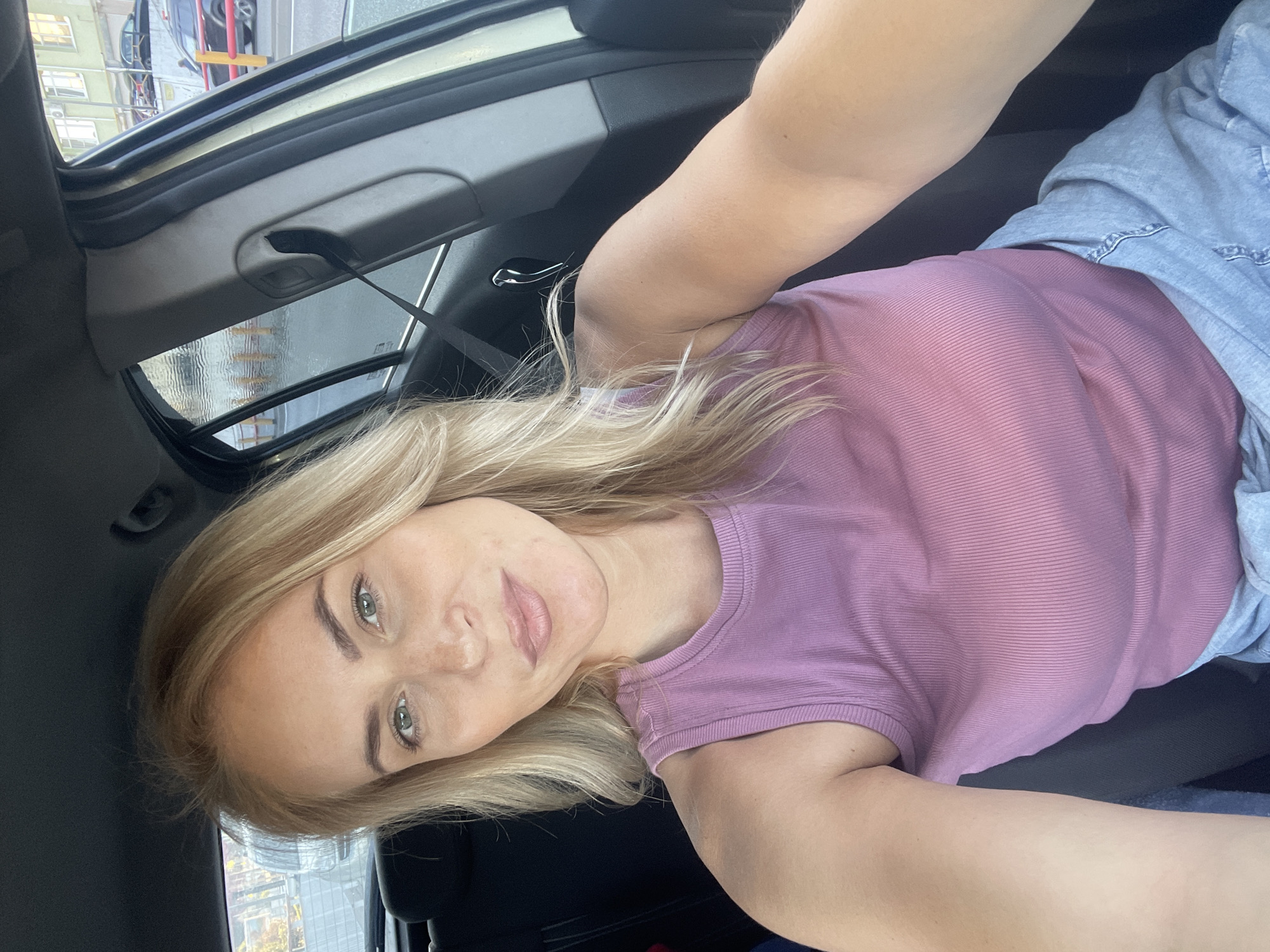 The image appears to be a selfie taken inside a car. A blonde woman wearing a pink top and a grey jacket is looking at the camera. The interior of the car is visible, including the dashboard and safety belt. Outside the car window, buildings and possibly other vehicles can be seen. There is not enough information provided to confirm the image was taken in Sochi, Russia.