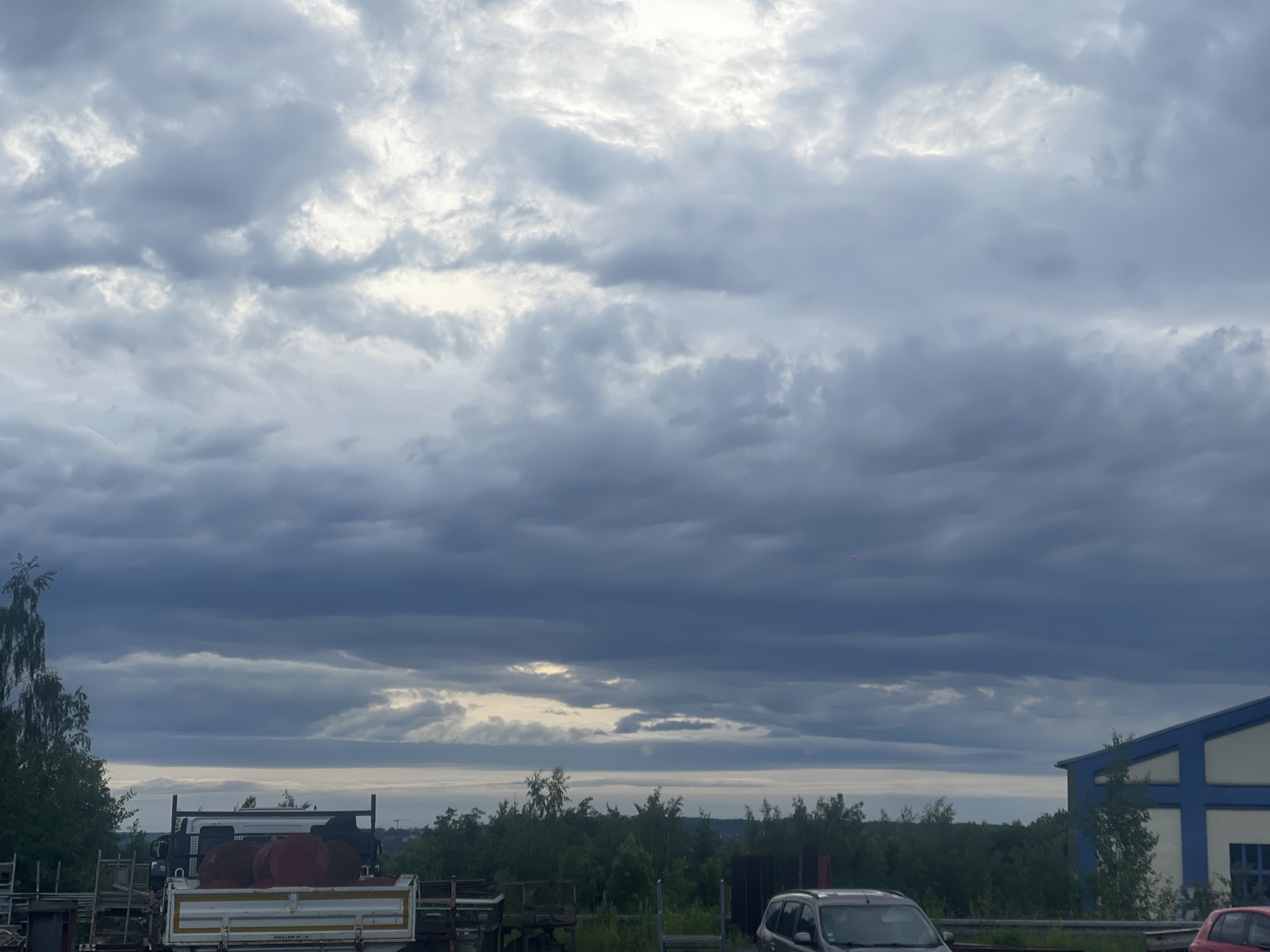 The image captures an overcast daytime scene, dominated by a heavy, cloudy sky. The clouds exhibit various shades of grey and white, with brighter patches suggesting diffused light breaking through. Towards the horizon, a band of lighter sky is visible, separating the dense clouds above from the distant landscape below, which appears to be either a flat landmass or a body of water.
In the mid-ground, a dense line of green trees and bushes stretches across the frame, obscuring any features beyond. On the far left, a few taller trees with lighter green foliage are visible.
The foreground features an industrial or commercial setting. On the left and center, various metal structures, possibly storage racks or scaffolding, are present. A white flatbed truck with a dark cab is parked, carrying large, reddish-brown cylindrical objects that resemble industrial barrels or spools. Partially visible text on the truck's bed reads "MEILLER KIE...". To the right, the front portion of a silver or grey car is parked, and a small part of a red car can be seen at the extreme right edge. Further right, a building with a blue lower section and white upper facade is visible.
A tiny, faint red dot is discernible in the upper right quadrant of the sky. The overall impression is a quiet, cloudy day in an area combining industrial elements with natural vegetation.
