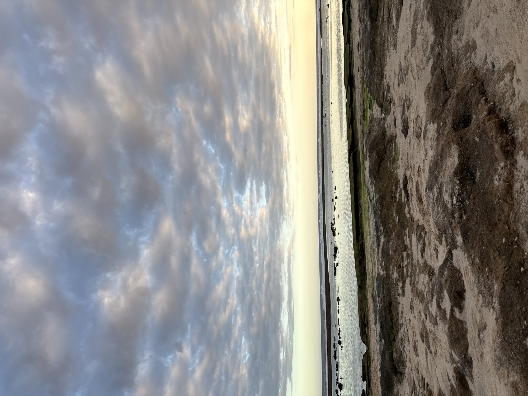 The image shows a beach scene at sunset. The left side of the frame is dominated by a sky filled with textured gray and white clouds, hinting at the setting sun. The clouds are illuminated with a soft, golden light. A narrow strip of water separates the sky from the beach. The water reflects the golden light of the sunset. The right side of the frame shows the sandy beach, with variations in color from light tan to dark brown. Some green algae or seaweed is visible on the rocks along the water's edge. The image is taken from a low angle, emphasizing the vastness of the sky and the texture of the beach. The location is likely in Kuta Selatan, Indonesia, given the tropical coastal environment. There are no people visible in the scene.