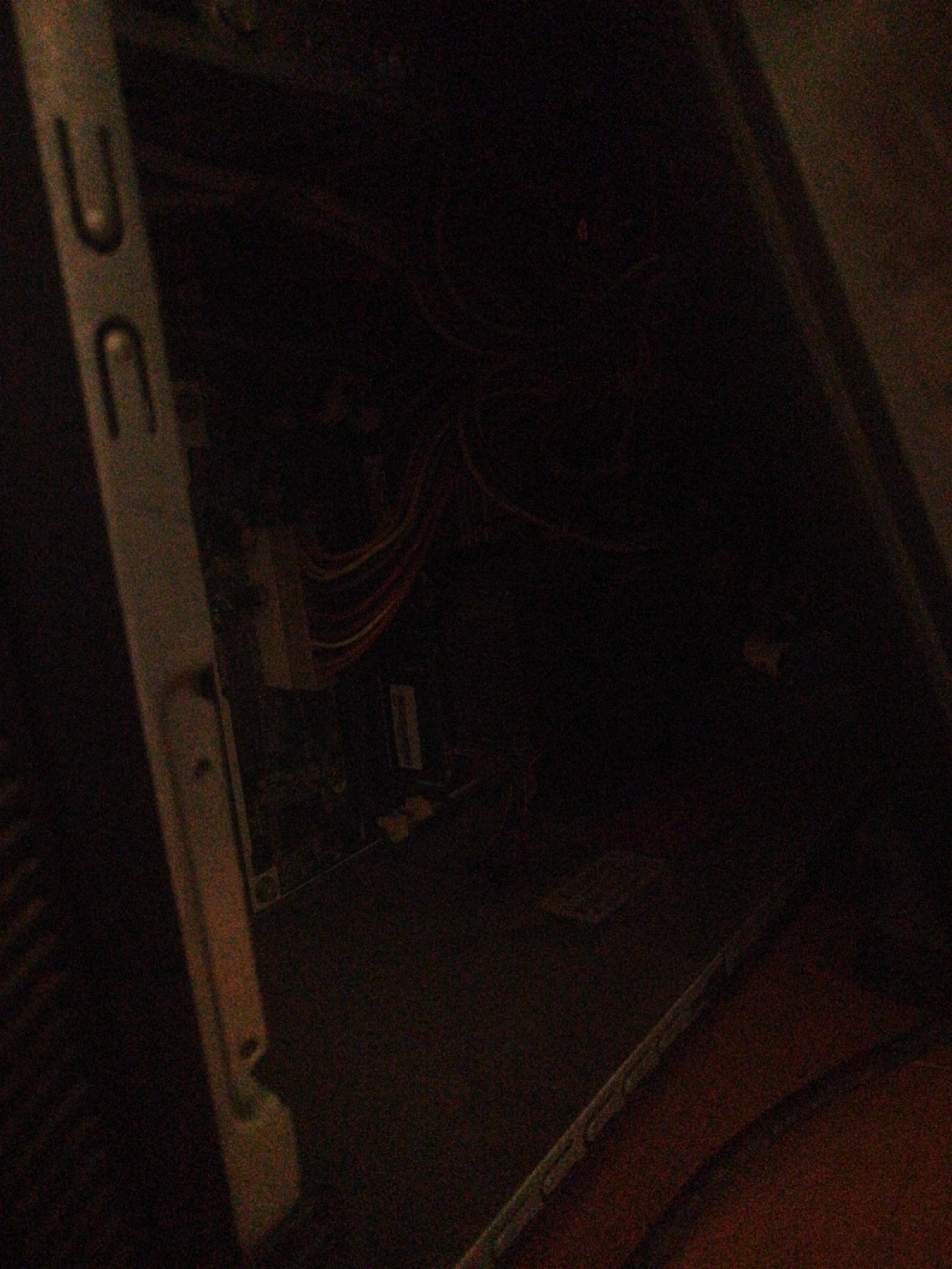 The image shows the interior of a dark computer case. A vertical metal bracket with two U-shaped openings is visible on the left side. The bracket is gray and shows signs of wear. To the right of the bracket, various wires, including red, white, and orange cables, are visible, leading to what appears to be computer components. The overall environment is dark, with the lighting primarily focused on the bracket and the visible wires. The background is obscured by shadow. There are no people or animals present in the image.