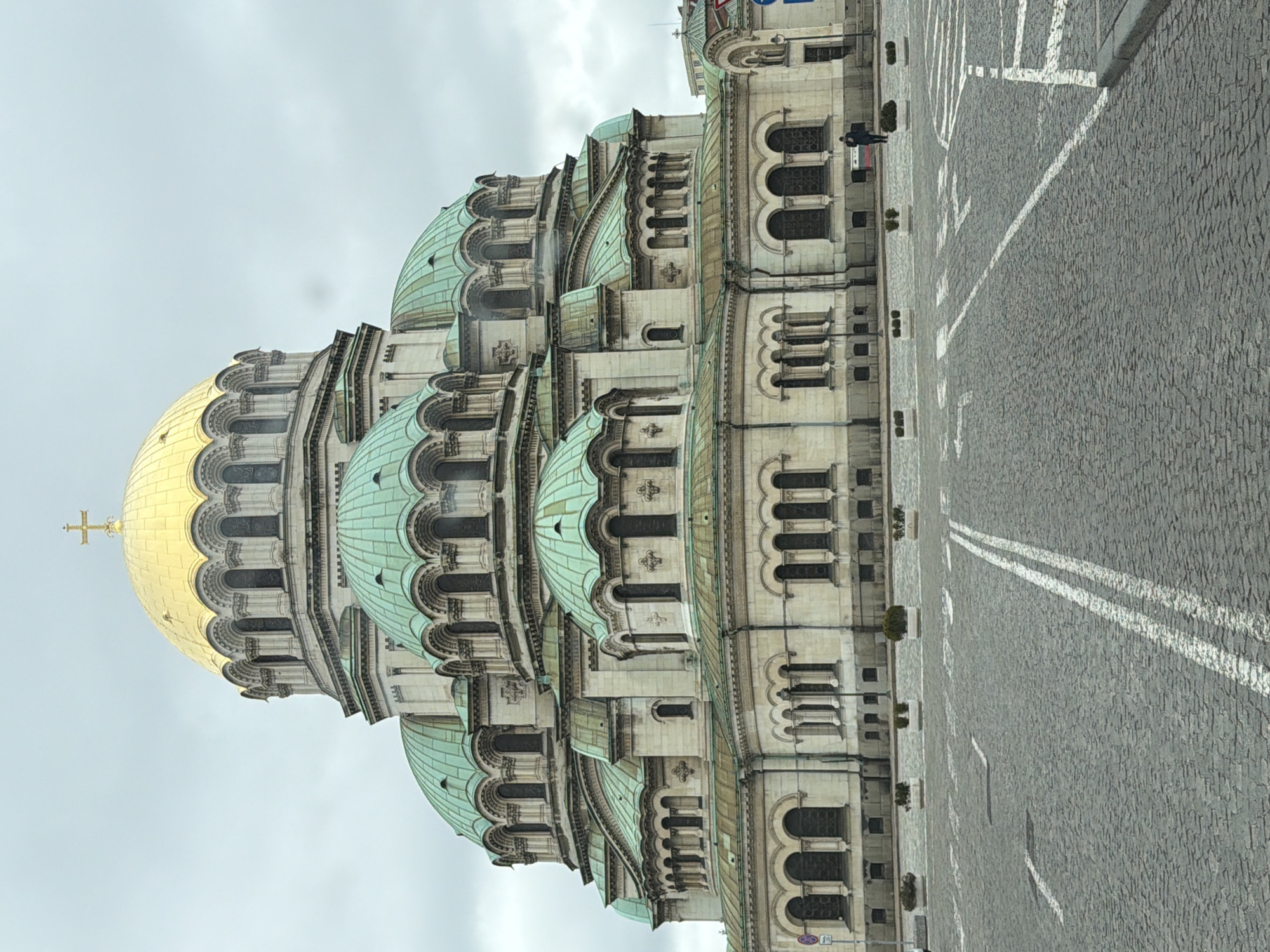 The image shows the Alexander Nevsky Cathedral in Sofia, Bulgaria. The cathedral has a large gold dome and multiple green-colored domes. The building is light-colored stone and features arched windows. A paved street runs along the side of the cathedral, with white lines marking lanes. A person in dark clothing stands on the sidewalk near a traffic sign. The sky is overcast.
