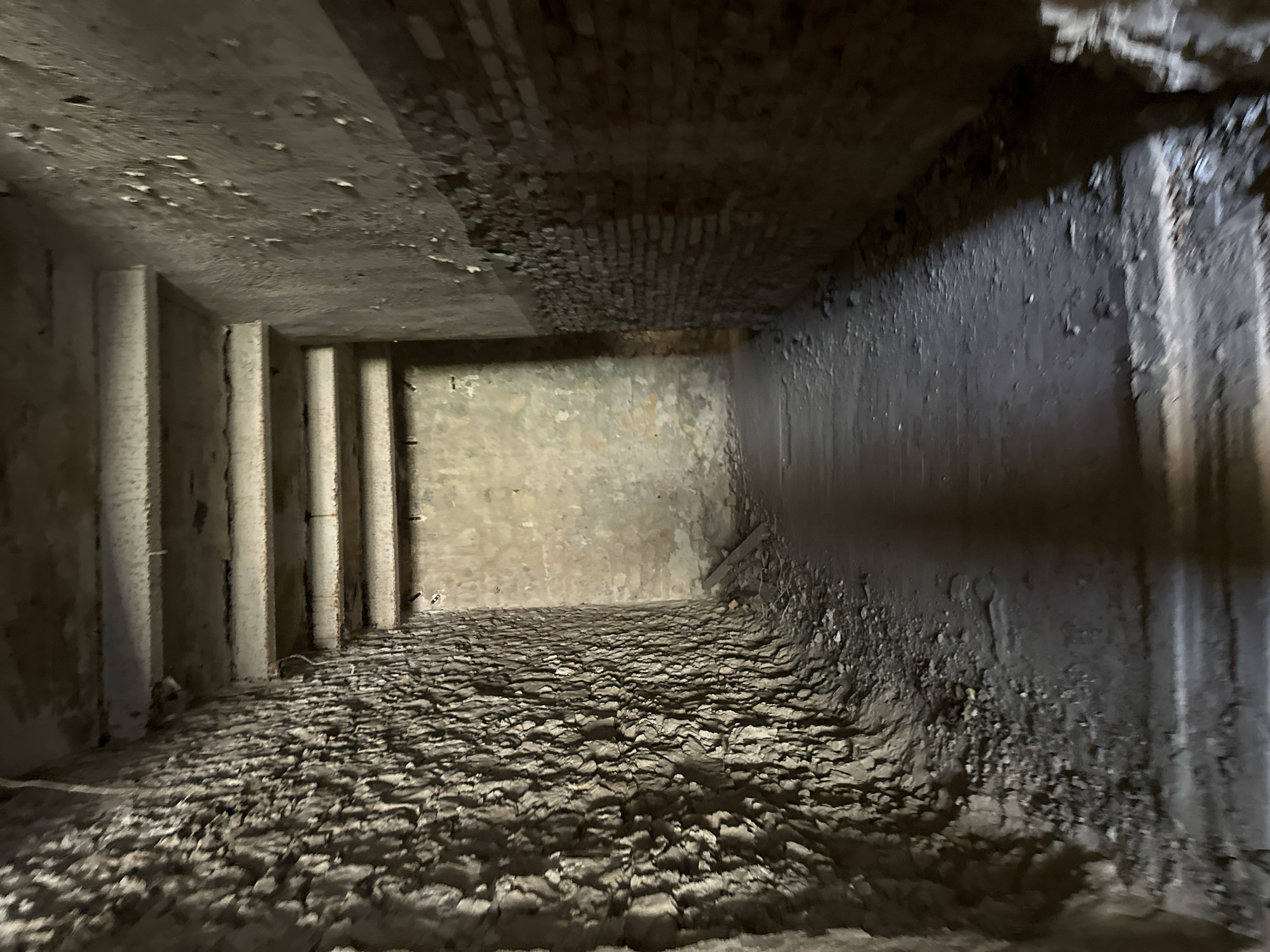 The image depicts an interior view of a dark, narrow corridor with textured walls and a rough floor. The walls on the left are a dark, rough stone, while the right wall transitions from brick to a rough, uneven plaster. The ceiling is composed of exposed concrete beams. The floor is covered in dirt and debris. The scene appears to be an abandoned or derelict structure, with no signs of human presence. The lighting is dim, casting shadows and highlighting the rough textures of the surfaces. The perspective suggests the viewer is standing within the corridor, looking forward into a slightly brighter, more open space. The environment is consistent with an underground structure or an old, neglected building. The location is identified as Abaata, Abkhazia.