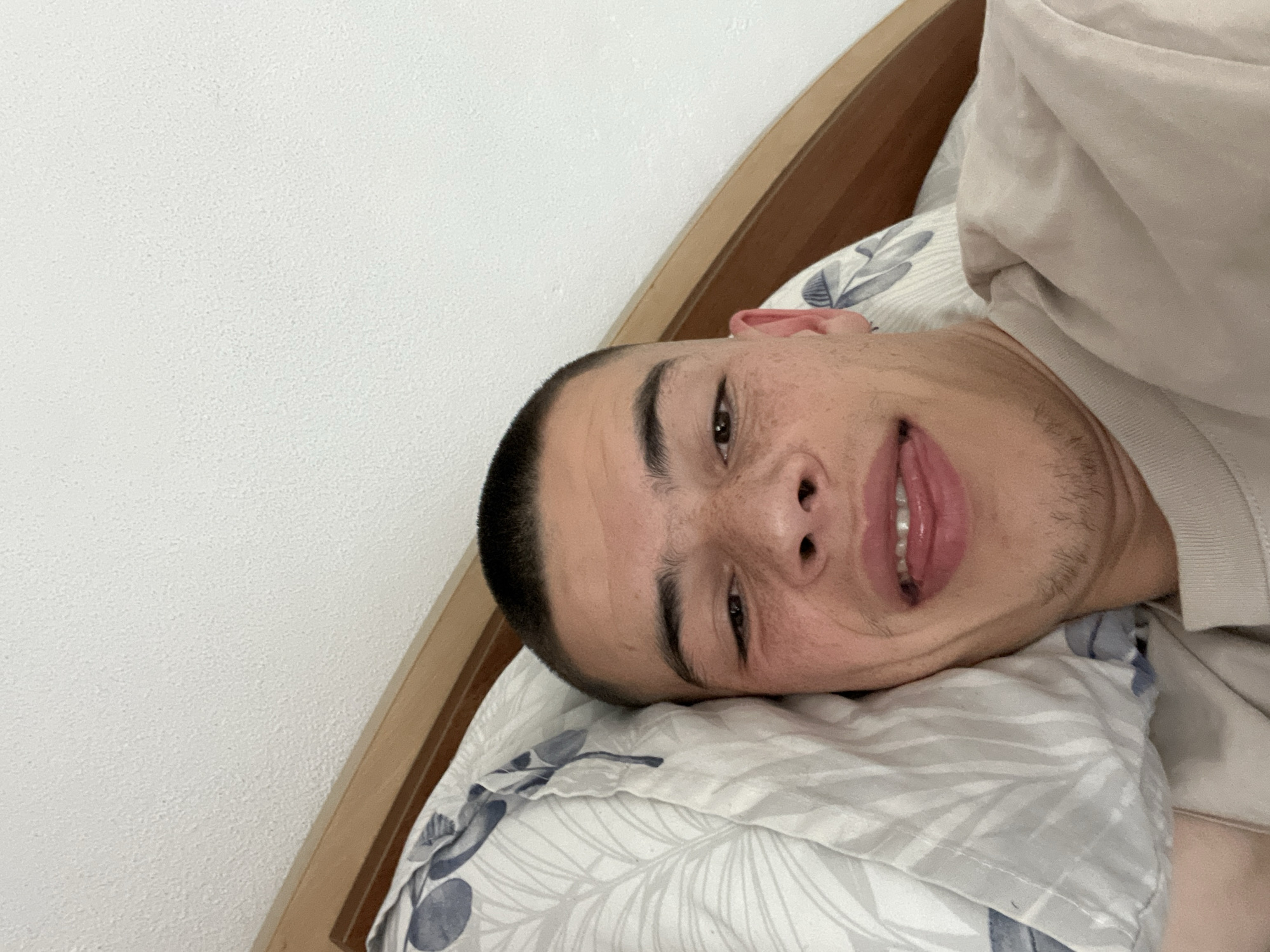 A young man lies on a pillow with a blue and white floral pattern, possibly in a bed, angled towards a white wall with a wooden headboard. He is on his side, close to the camera, with a short haircut and light stubble. His mouth is slightly open, revealing his teeth. The location in Karavelovo, Bulgaria, is not verifiable from the visual information provided.