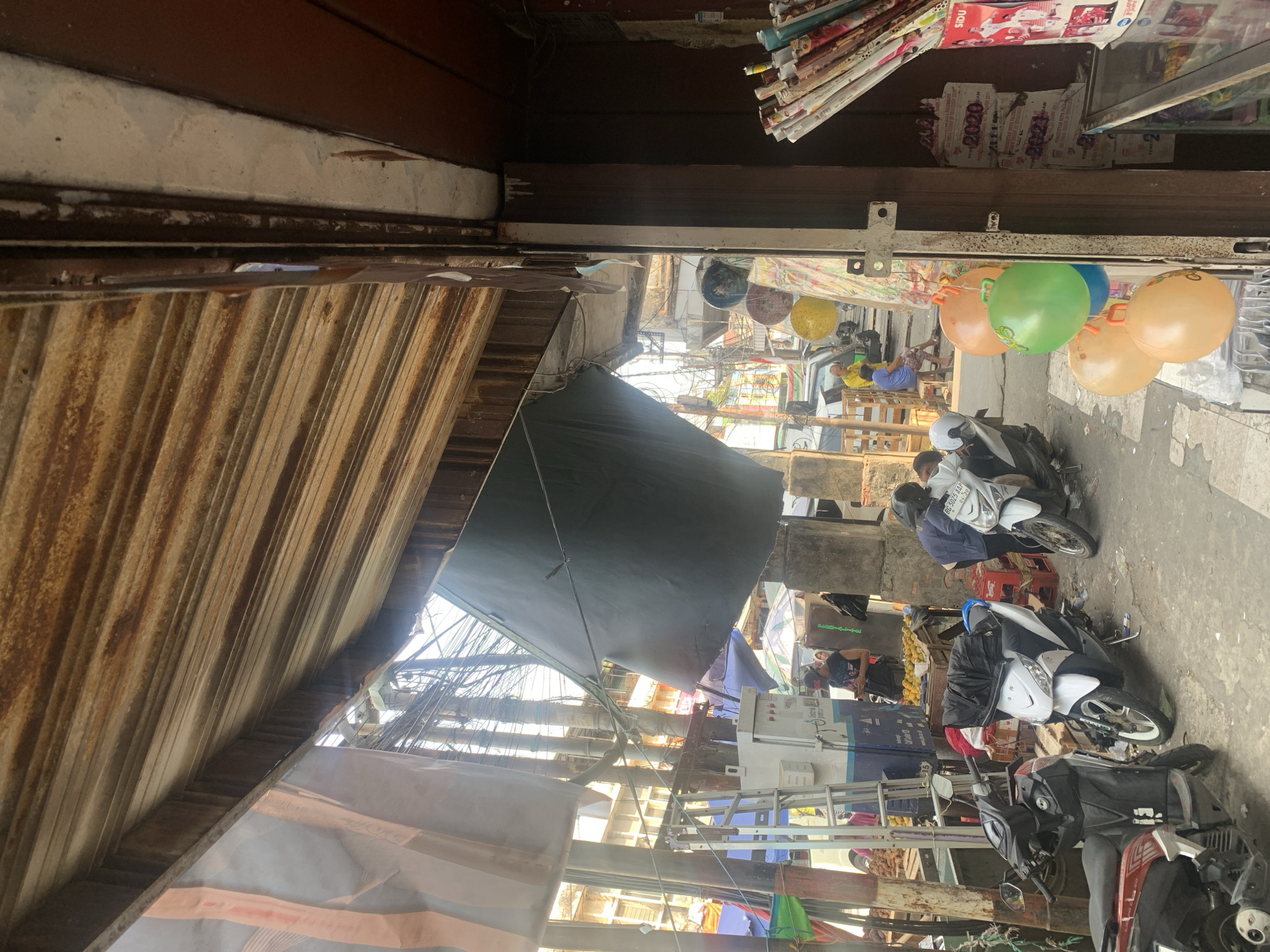 This image captures a street scene in Palembang, Indonesia, likely a market area, characterized by a makeshift outdoor stall. A corrugated metal roof, showing signs of rust and wear, extends over the scene. In the foreground, several motorbikes are parked. A dark-colored motorbike is on the left, followed by a white motorbike in the center, and another white motorbike to the right.
Behind the motorbikes, a street vendor's stall is set up. There are stacks of oranges on a table. Several people are visible around the stall; one person in a blue shirt is interacting with the vendor, and another person in a black tank top and baseball cap is looking at their phone. In the background, more stalls and buildings are visible, along with a network of electrical wires suspended overhead.
Hanging from the stall are colorful balloons, possibly for sale, and rolls of patterned paper or fabric. A white electrical box with blue accents and a ladder are positioned next to the stall. The overall environment suggests a busy, informal commercial setting with typical urban street elements.