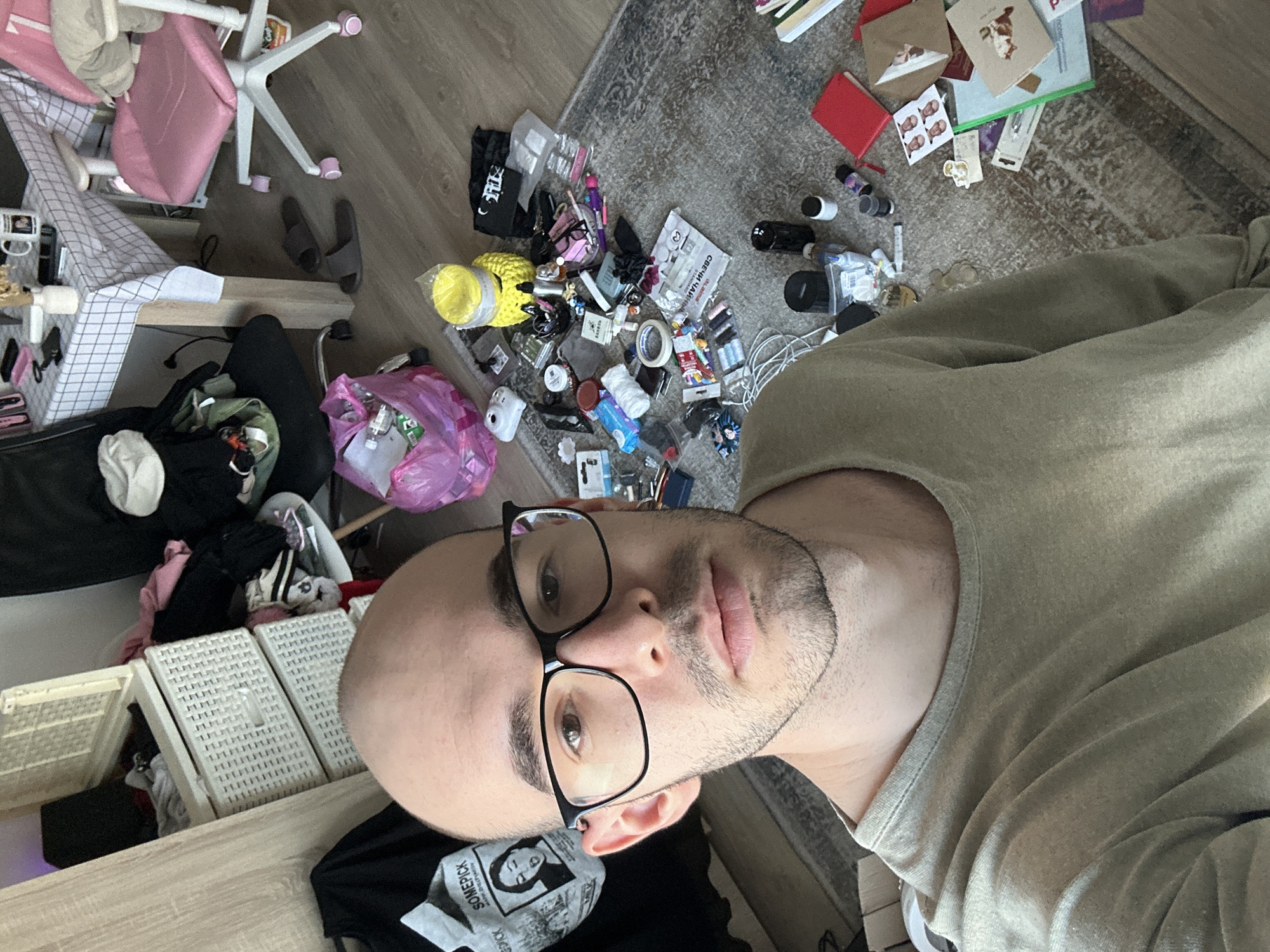 The image shows a man with a shaved head and glasses, wearing a light green sweatshirt. He is indoors, in what appears to be a cluttered room. To his left, there is a desk with white plastic storage bins, a pink chair, and a pair of gray slippers on the floor. In front of him, there is a pile of assorted items including a yellow yarn ball, a pink plastic bag, and various cosmetic and electronic items scattered on the floor. In the background, there is a rug and additional scattered objects. The location of the image is not explicitly stated. However, the objects found in the image do not provide any clues regarding Lyubertsy, Russia.