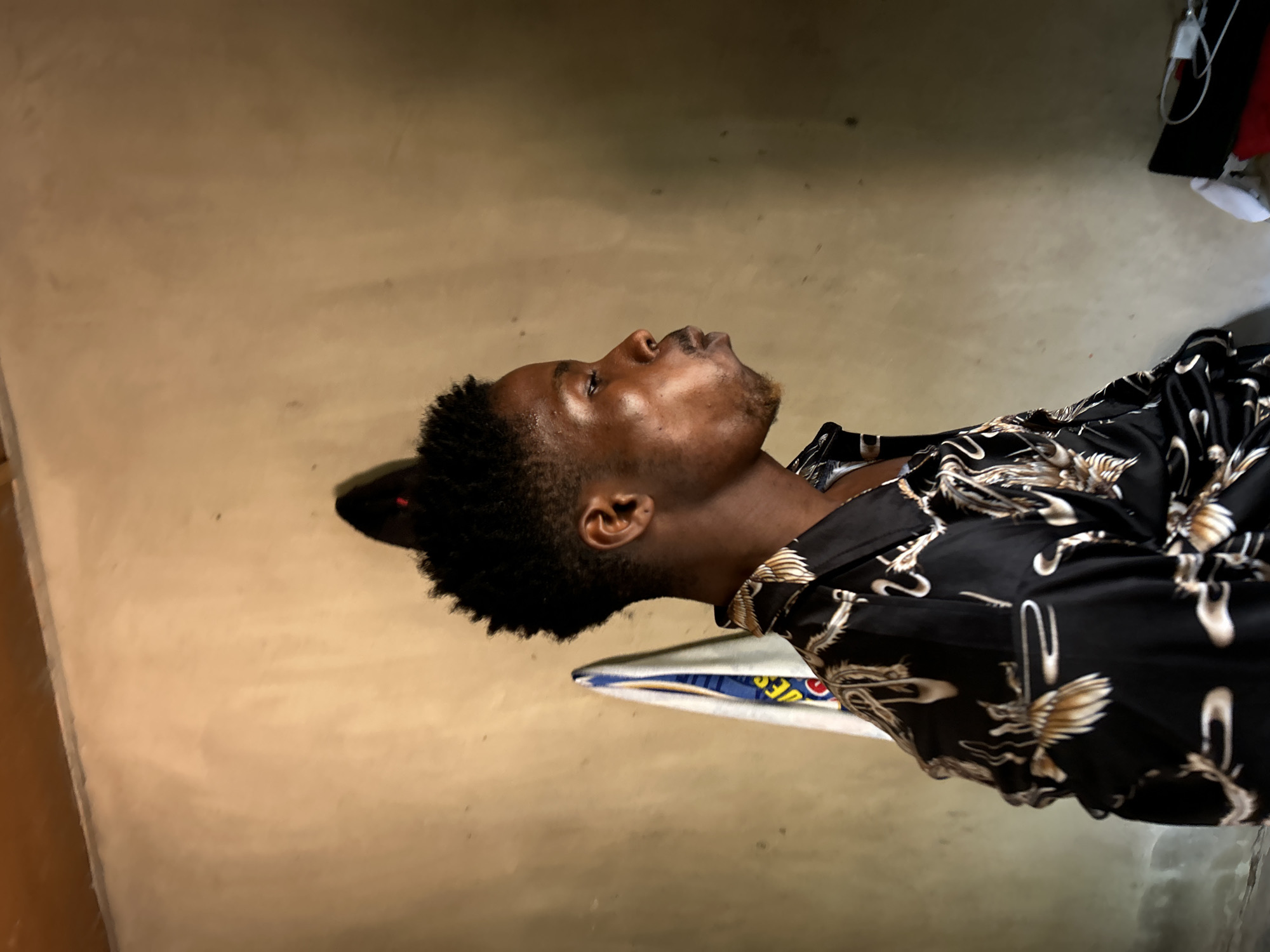 A man is laying down. He has short, dark hair and is wearing a black shirt with a crane-like bird pattern. The location is a room with a beige wall and floor.