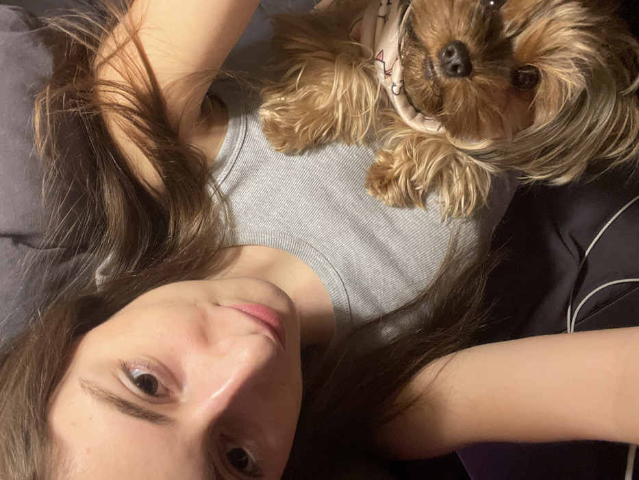 A woman with light skin and brown hair lies on a gray surface, gazing into the camera. A Yorkshire Terrier wearing a patterned garment rests on her chest. The woman's hair is spread around her head on a gray pillow or surface. The dog faces the camera, and its paws are visible. The woman's arm is visible on the right side of the frame, against a dark background, potentially clothing. The lighting appears soft, highlighting the subjects.