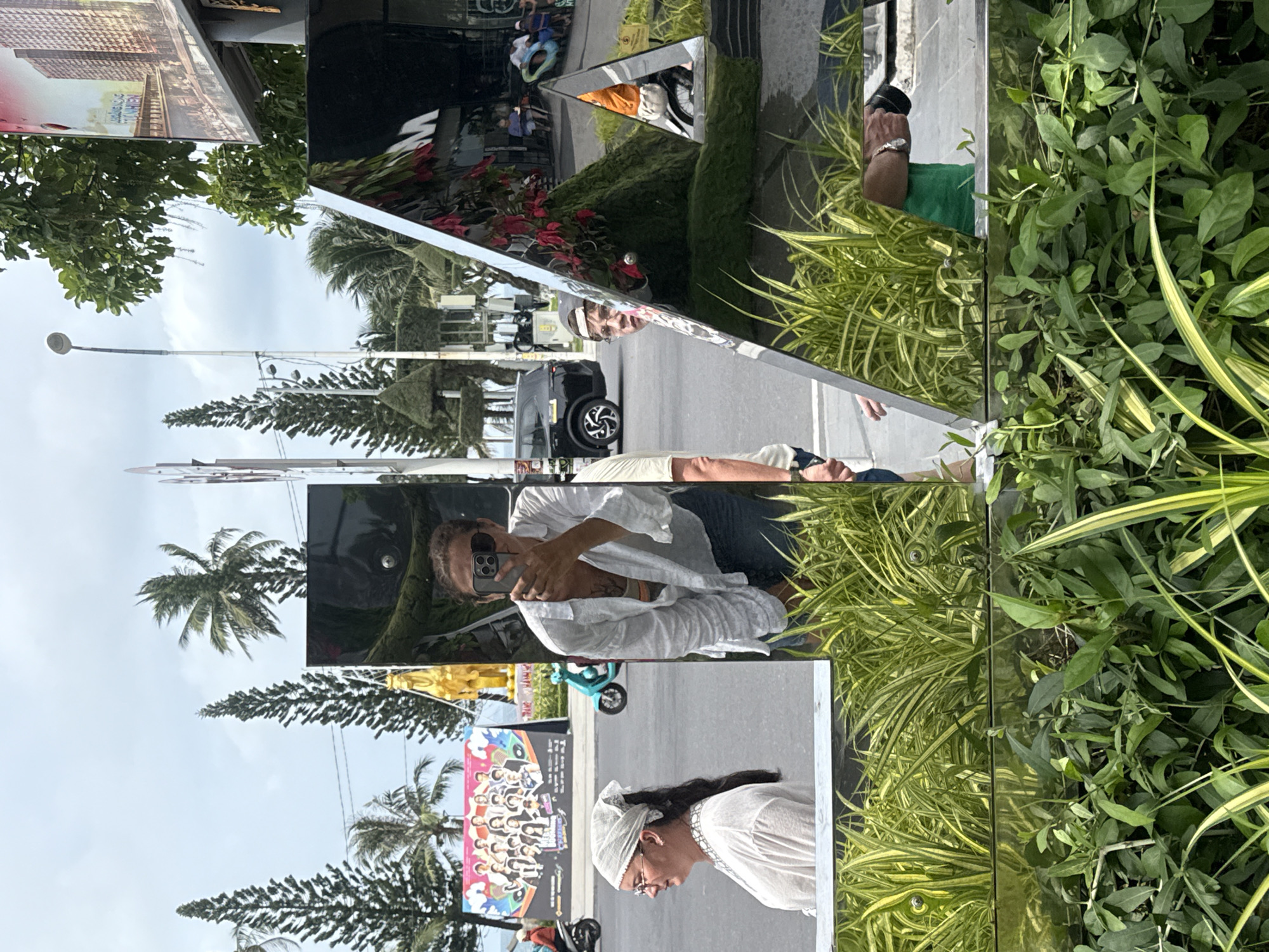 The image captures a street scene in Nha Trang Ward, Vietnam, as reflected in multiple mirrored surfaces. In the foreground, lush green plants with striped leaves are prominently featured, with some appearing to be in planters. In the background, a street with a roadway and sidewalk is visible.
Multiple reflections are observed:
* A woman wearing a white headscarf and glasses is reflected in a large, flat mirror, looking to the left.
* Another woman, wearing a white shirt and sunglasses, is taking a selfie with a phone in a vertical mirror. She has a tattoo on her chest.
* A young person wearing glasses and a baseball cap is reflected in a triangular mirrored sculpture, looking towards the viewer.
* A person wearing a green shirt is reflected in the same triangular sculpture, holding a black object, possibly a camera or phone.
* Further reflections show buildings, trees (including palm trees and tall, conical trees), a poster with a group of people on it, a gold statue, a turquoise scooter, a black SUV, and other pedestrians and vehicles. The overall environment appears to be a public area with commercial establishments and thoroughfares.