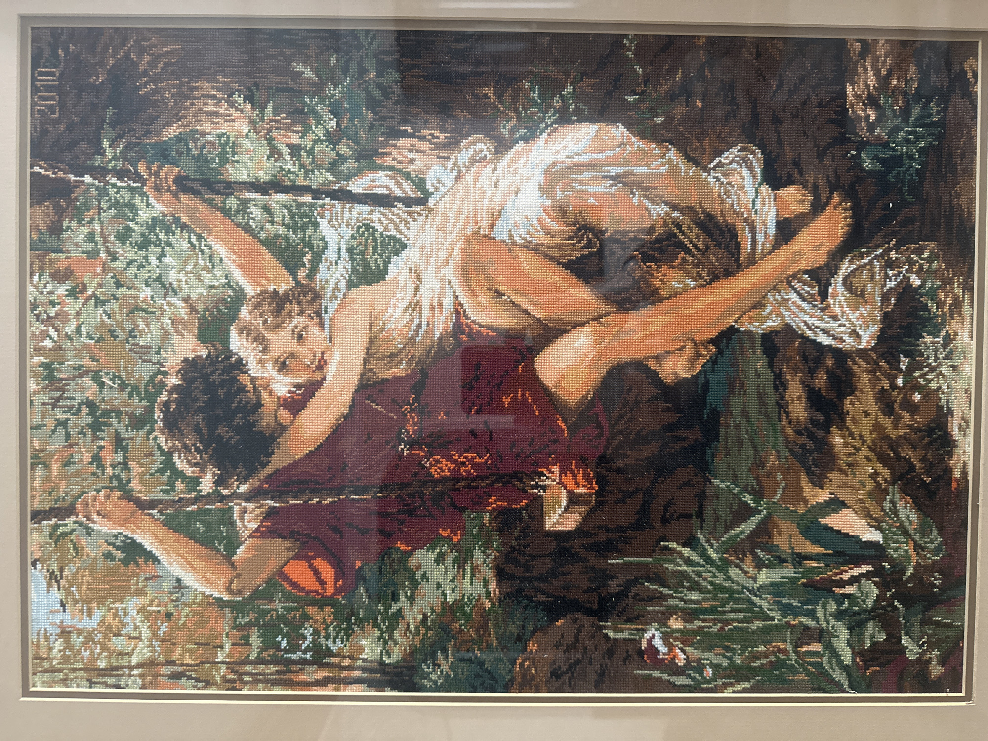 This media file is a framed cross-stitch artwork depicting a scene of two people, a man and a woman, in a close embrace. They appear to be lying down, possibly on a swing or hammock, amidst lush greenery. The man is positioned below the woman, his arms around her. The woman is looking towards the viewer with a gentle expression. Both figures are rendered with a warm, earthy color palette, with the woman draped in flowing, light-colored fabric. The background is filled with various shades of green and brown, suggesting a natural, outdoor setting, possibly a forest or garden. The overall mood is intimate and serene. In the upper left corner of the artwork, the year "2010" is visible, embroidered in dark thread. The artwork is housed in a simple, light-colored frame. The file was likely taken in Moscow, Russia.