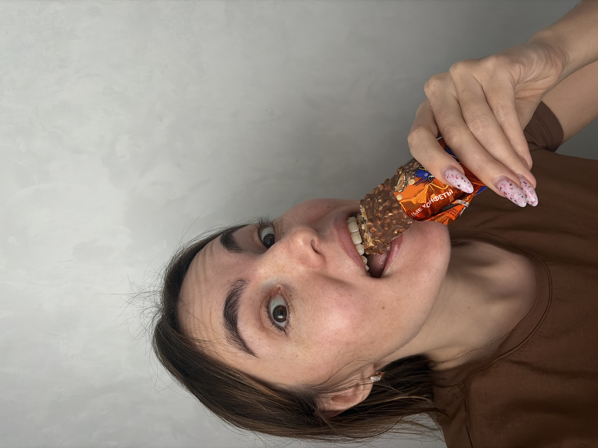A person is holding an unwrapped chocolate bar in their mouth. The person is wearing a brown t-shirt. The chocolate bar has a brown and orange wrapper. The nails of the person are long and decorated with pink patterns. The background is a gray wall. There is no evidence of Neftekamsk, Russia.