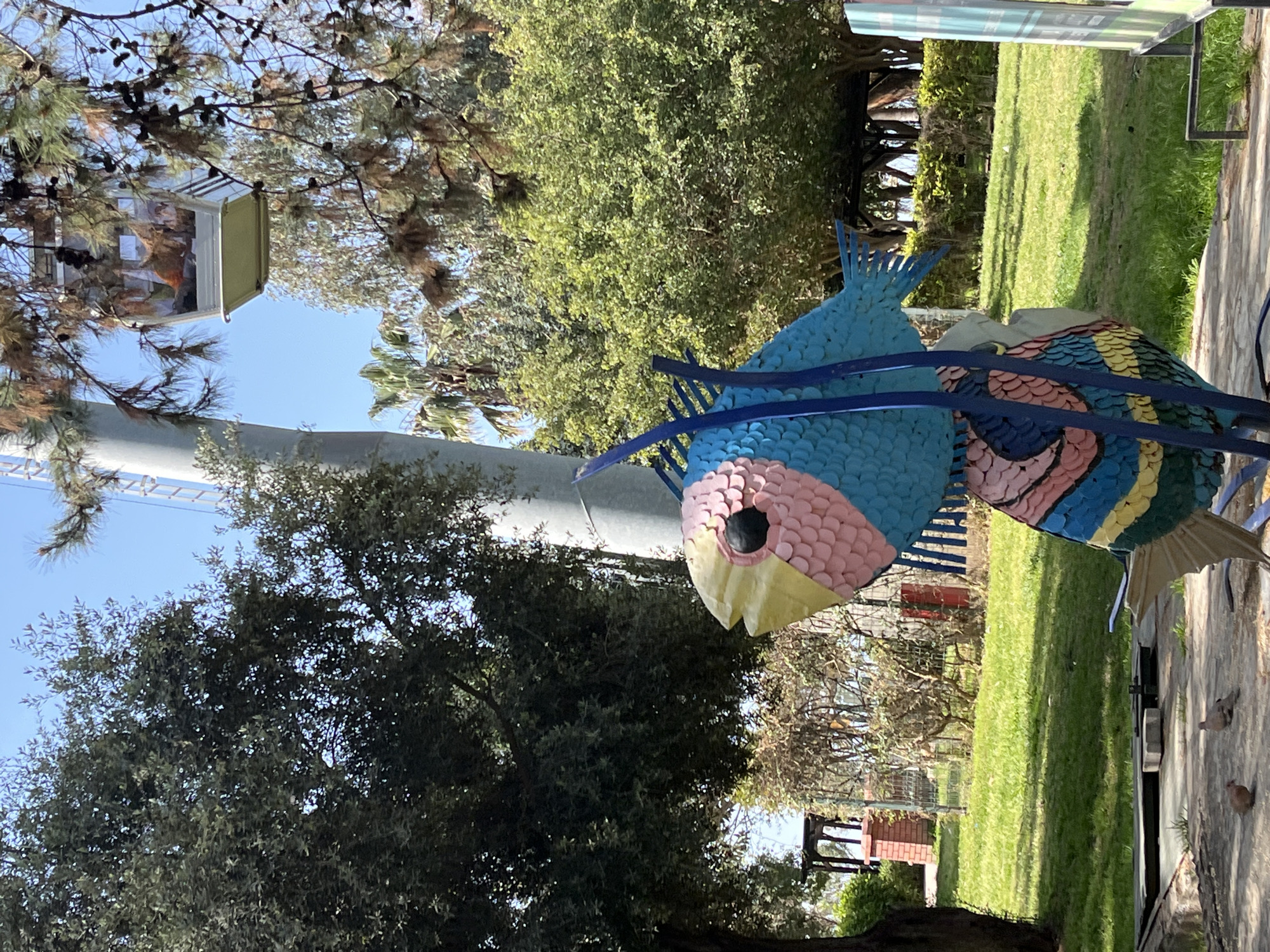 The image features a colorful fish sculpture made of bottle caps in Alanya, Turkey. The fish is predominantly blue with pink and yellow details on its head. Another fish sculpture, partially visible, displays blue, pink, and yellow patterns. They are constructed with blue metal supports.
The sculptures are situated in a park or garden, as indicated by the lush green grass and trees in the background. A tall, white structure (possibly a pillar or tower) is visible in the background. The sky is clear and blue.