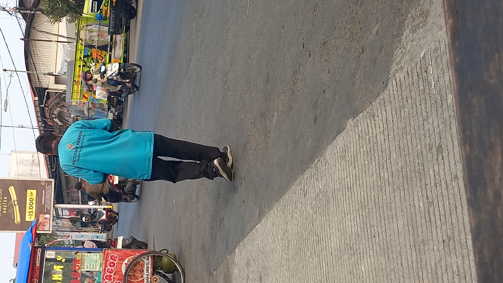 A medium shot captures a street scene in Mangunsari, Indonesia, featuring a parking attendant in a bright blue shirt walking away from the camera. The attendant wears dark pants and athletic shoes. In the background, a food cart with a red and white striped awning and a large wheel is visible on the left. Several motorcycles are parked or in motion on the street, and a colorful truck with "MITSUBISHI" branding is on the right. The environment appears to be a commercial or busy street area, with buildings and advertisements lining the road. The asphalt street has a textured concrete border on the left side. The lighting suggests it is daytime.