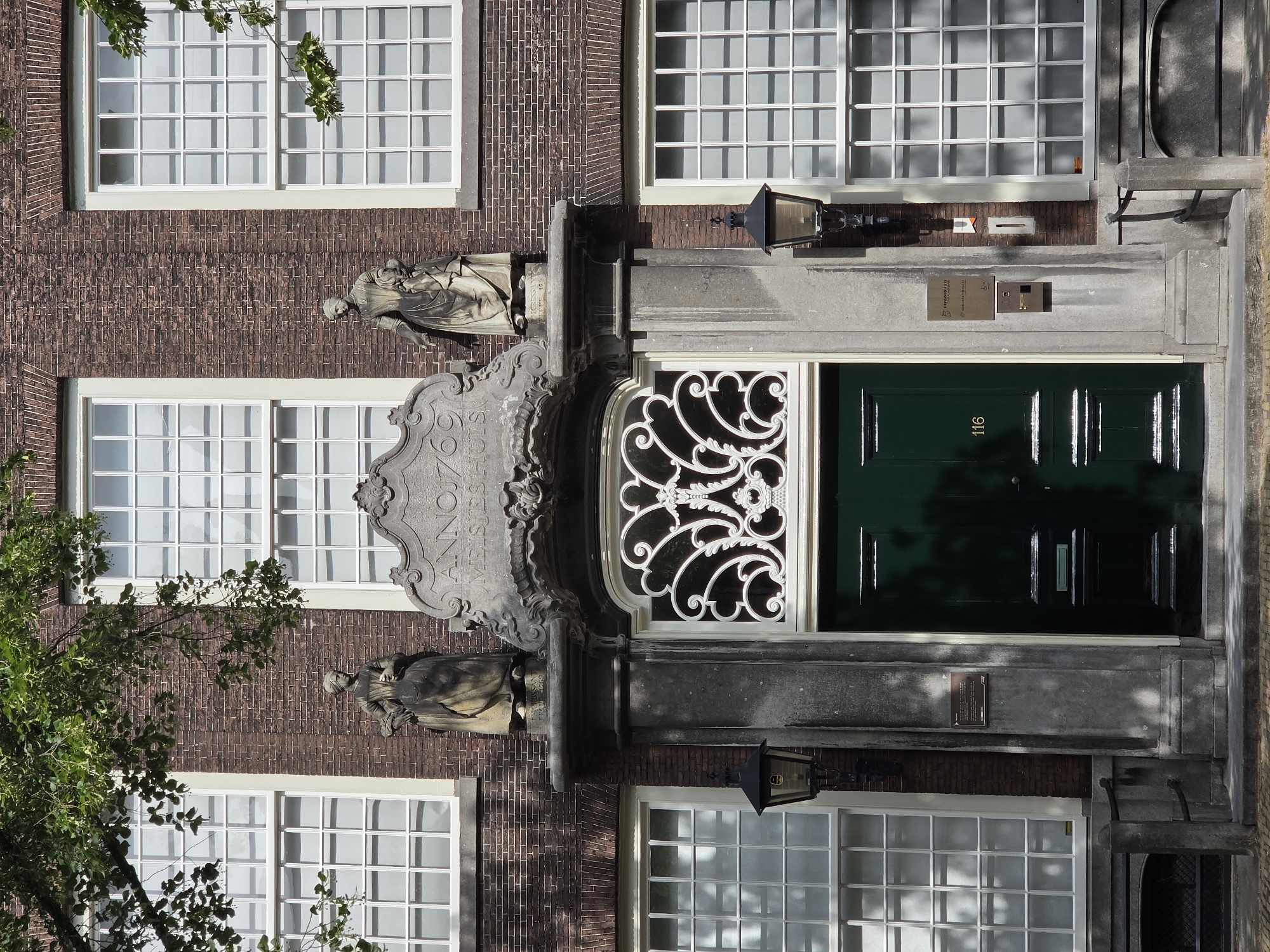 The image shows the exterior of a historic building in Delft, Netherlands. The building is made of brick with large white-paned windows. Above the entrance is a stone carving with the inscription "ANNO 1769 MEISJESHUIS". Two stone statues flank the inscription. The entrance itself features a dark green door with ornate metalwork above it. A street lamp is visible to the left of the door, and a metal railing runs along the base of the building. A small plaque is mounted on the wall to the right of the door. Trees with green leaves overhang the top of the building, casting shadows on the facade. The scene appears to be a still, daytime shot of the building's facade.
