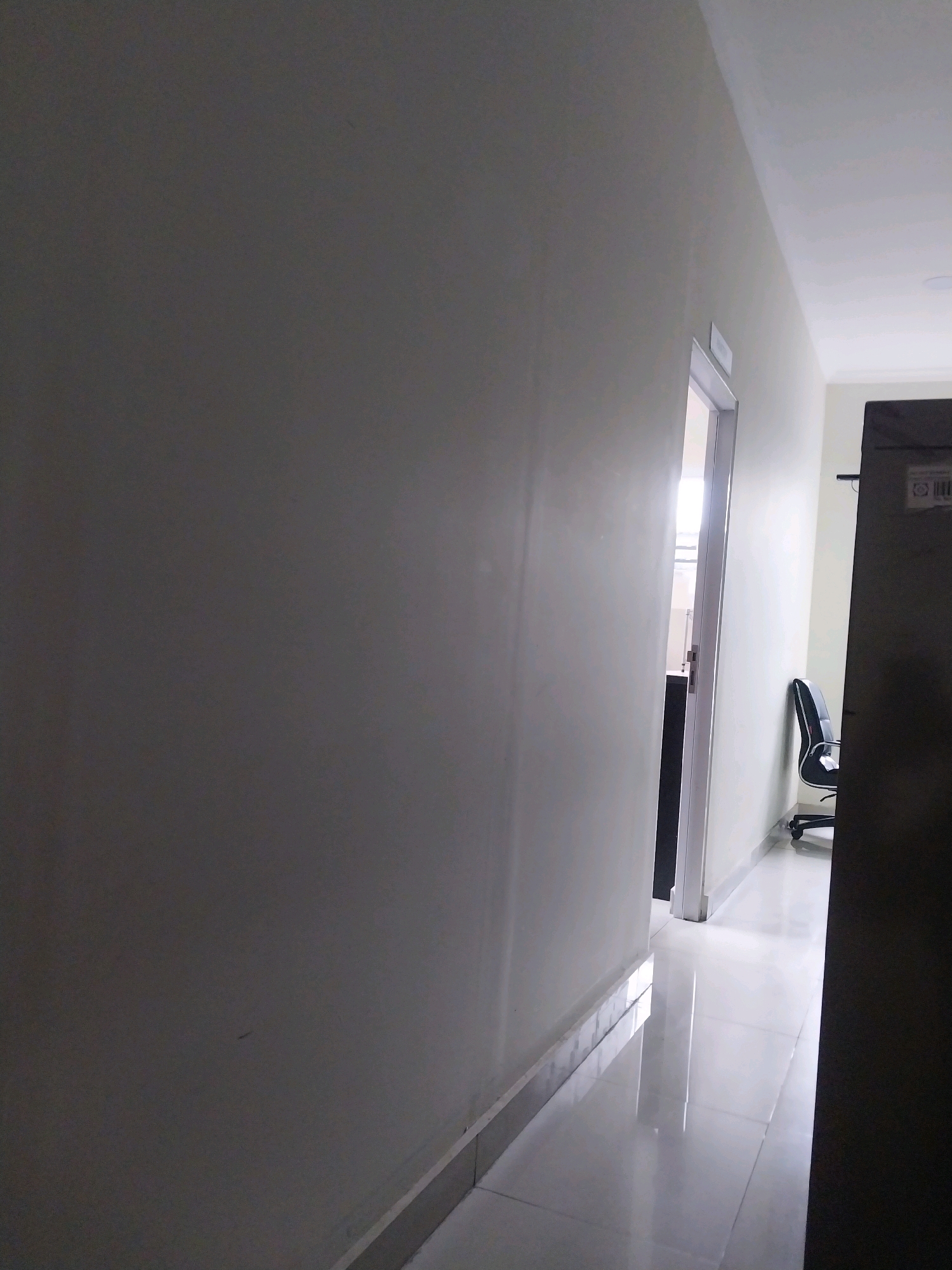 A dimly lit interior corridor shows a wall on the left and a door opening into another room on the right. The floor is tiled and reflects light. A black office chair with wheels is visible in the adjacent room. The scene appears to be in Sleman, Indonesia.