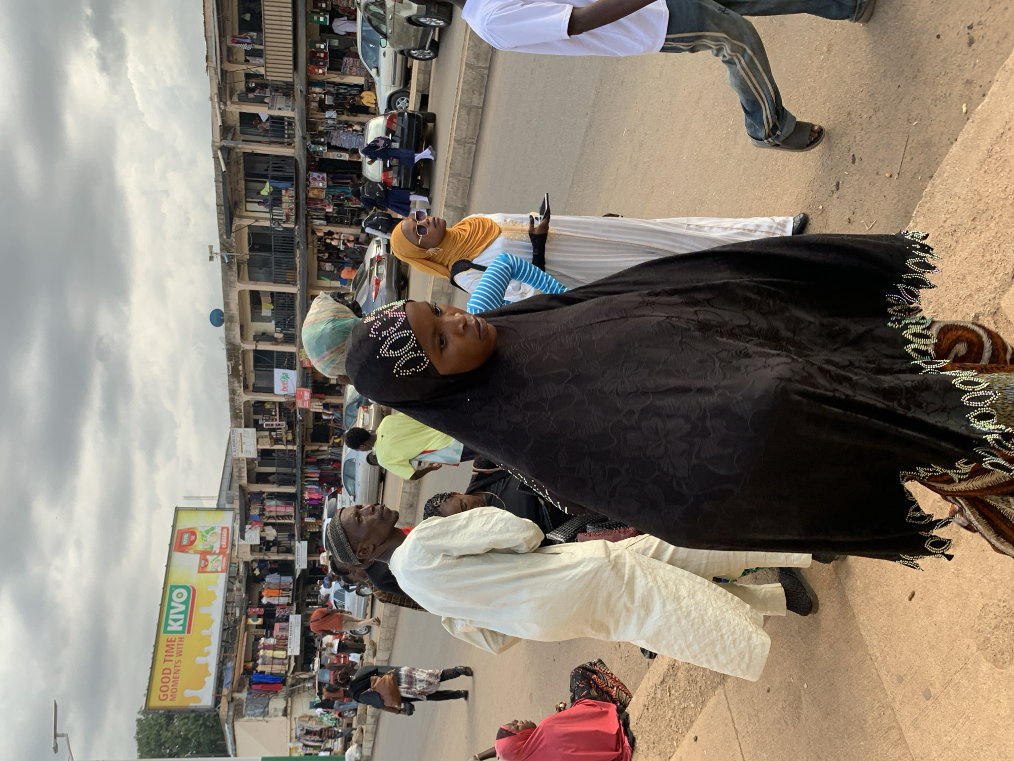 The image captures a bustling street scene, likely in Jos, Nigeria, with a prominent marketplace and street activity. In the foreground, a woman is dressed in a black hijab with intricate beadwork around the neckline and hem. To her right, another woman wears a yellow hijab and a white garment, holding a smartphone. Behind them, a man in a cream-colored robe and skullcap looks upwards.
The background reveals a two-story building filled with shops, showcasing a variety of goods. Cars are parked along the street, and several other individuals are visible, some walking and others browsing the shops. A large billboard with the text "GOOD TIME MOMENTS WITH KIVO" and images of a Kivo product is mounted on the building. The sky is overcast, suggesting a cloudy day. The overall impression is one of a busy commercial area with people engaged in daily activities.