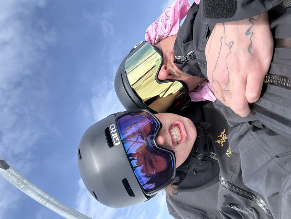 Two individuals are depicted in a close-up, elevated outdoor setting in Le Monêtier-les-Bains, France. The scene is illuminated by a bright sky featuring blue expanse with sparse white clouds. In the upper left quadrant, a metallic gray structural element, consistent with a ski lift component, is partially visible.
The subject on the left wears a dark gray Giro-brand ski helmet and ski goggles with blue and purple mirrored lenses. Their mouth is open, revealing teeth and clear dental aligners. The goggles reflect a distorted image of a face and an indistinct environment. This individual wears a dark gray or black ski jacket with a visible Arc'teryx logo on the chest.
The subject on the right also wears a dark ski helmet and goggles, which have yellow and gold mirrored lenses. A pink patterned bandana is visible beneath their helmet and around their neck. A portion of their face, including a mustache, is visible. Their right hand, adorned with black line tattoos, is positioned on their dark ski jacket. The subjects appear to be in close proximity, suggesting they are seated together.