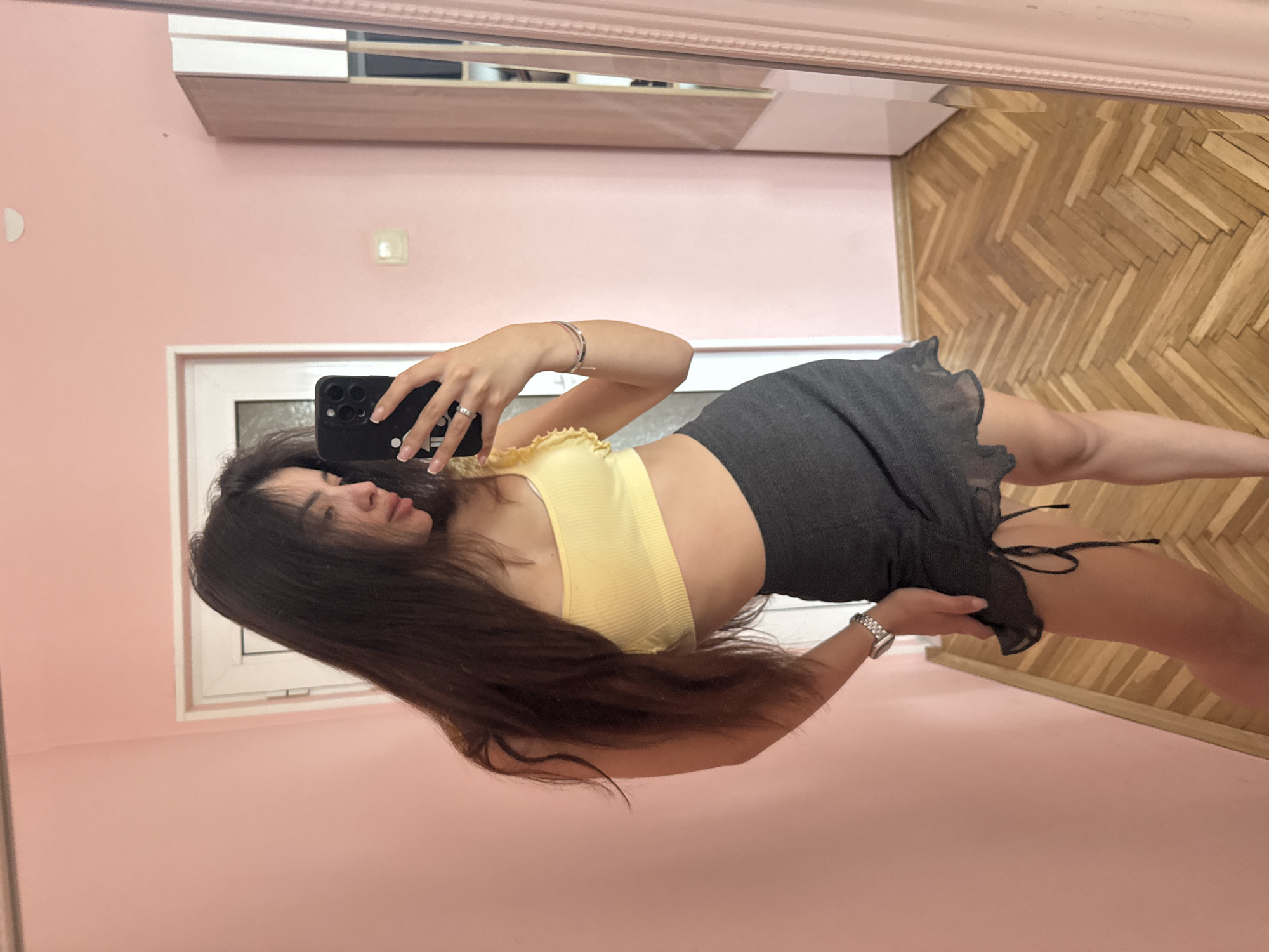 An indoor mirror selfie captures a young woman standing in a room. The image is rotated 90 degrees clockwise. She is holding a dark-colored smartphone with her right hand, its camera lenses visible, pointed towards the mirror to take the photo. Her left arm is bent at the elbow, and she wears a silver-toned watch on her left wrist and rings on her right hand. Her nails are manicured with light polish.
The woman has long, dark brown hair that falls over her shoulders and back. She is wearing a light yellow, ribbed crop top with ruffled trim along the neckline, and a dark grey or black mini skirt with ruffled detailing and a side tie. Her face is partially visible in profile, looking towards the phone, with a neutral expression.
The setting appears to be a residential room. The wall reflected behind her is painted a soft pink or peach color. A white-framed window is visible in the reflection behind her left shoulder. The floor, also reflected in the mirror, is made of light-toned wooden parquet in a herringbone pattern. Above the mirror's reflection of the pink wall, there is a reflection of what appears to be a light-colored wall with shelves or a cabinet. The mirror itself has a decorative, possibly pink-toned, frame. There are no specific location cues visible in the image to indicate Plovdiv, Bulgaria, and no visible text. The lighting suggests an indoor environment.