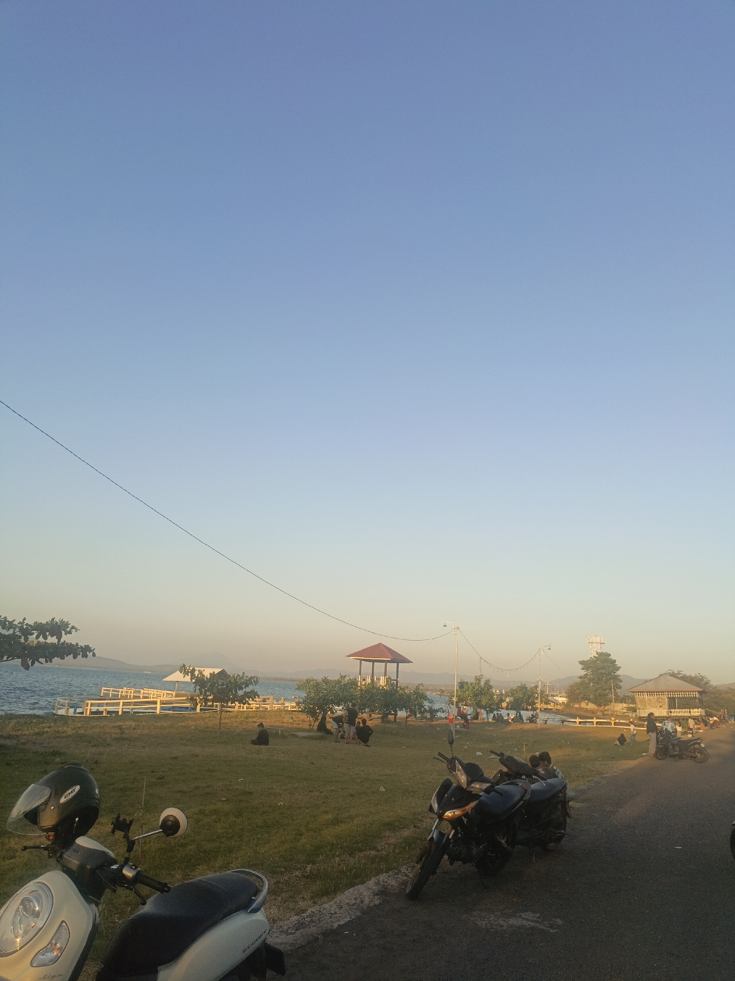 The image captures a waterfront scene in Lewoleba, Indonesia, during what appears to be late afternoon or early evening. The sky is a clear, gradient blue, transitioning to a lighter hue near the horizon. In the foreground, a white scooter is parked on the left, and to its right, two black motorcycles are parked on an asphalt road. The road runs alongside a grassy area where several people are seen sitting and standing.
Further back, a wooden pier or boardwalk with white railings extends into the calm blue water. On the water, there are a few small white structures, possibly gazebos or shelters, and a distant vessel. Several trees with green foliage are scattered throughout the park-like area. Two gazebo-like structures are visible in the middle ground: one with a red roof on the left, and another larger, open-sided building with a brown roof on the right. String lights are suspended between poles in the background, suggesting a recreational area. In the far distance, hazy mountains are visible. The overall atmosphere suggests a leisurely gathering at a public space by the sea.