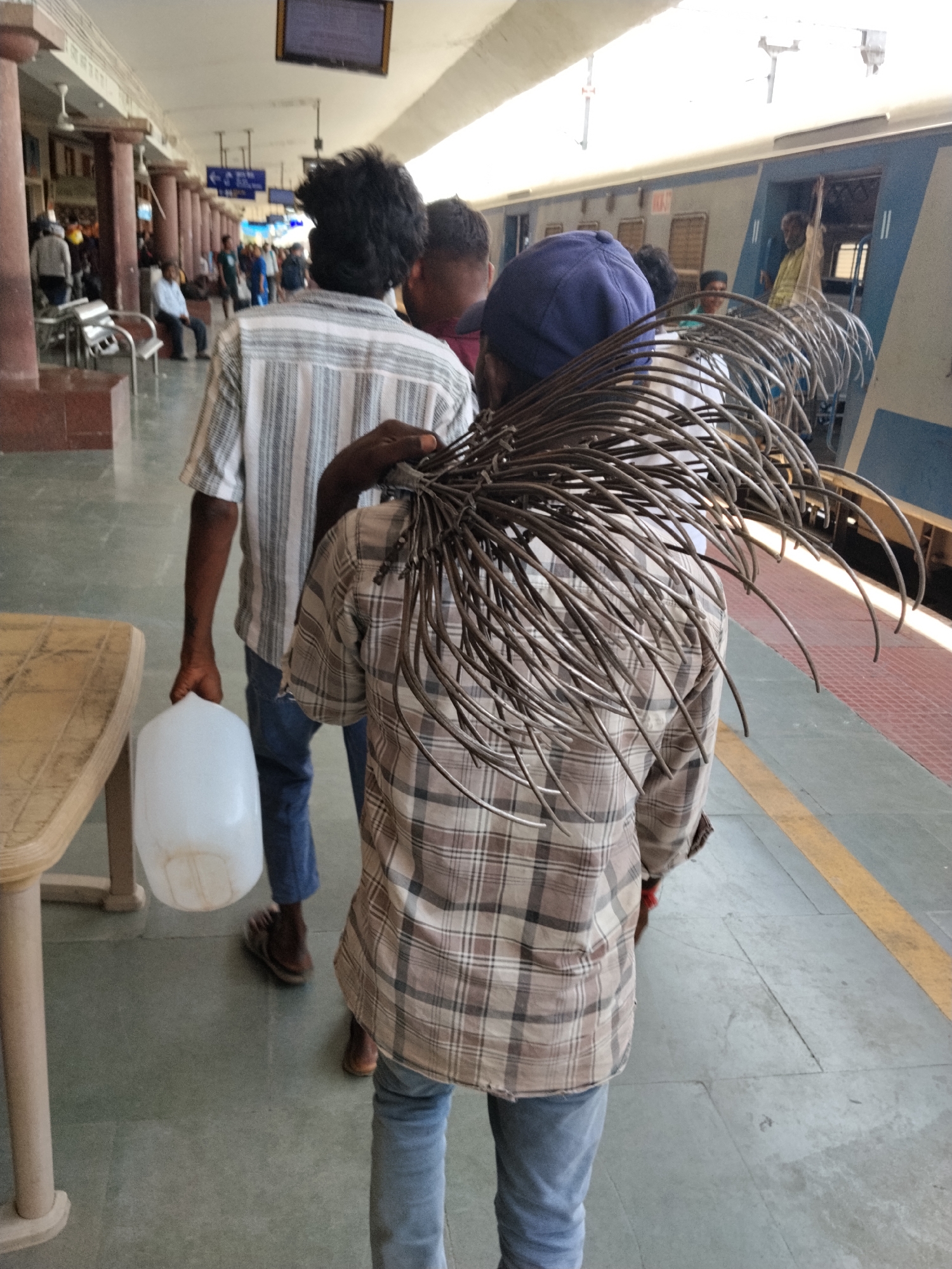 A man with a bundle of metal rods on his back walks through a train station in Mamillakunta, India. The man is wearing a plaid shirt, blue jeans, and a blue cap. Behind him is another man carrying a white plastic jug and wearing a vertically striped shirt and blue jeans. The train platform has gray tile flooring with a yellow line. A train is visible on the right, and people are seated in the background on the left.