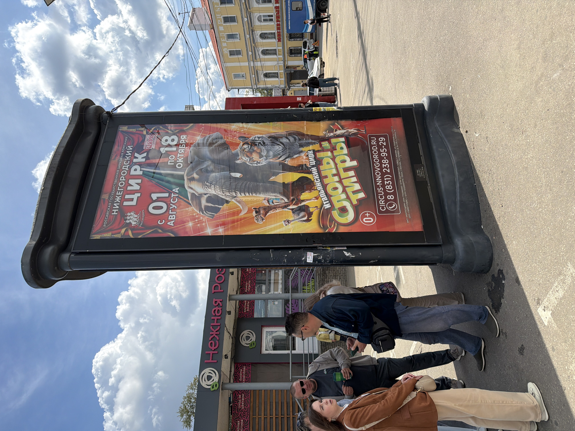 A group of people stand on a paved area near a large, double-sided circus advertisement in Nizhny Novgorod, Russia. The advertisement is predominantly red and features images of an elephant, a tiger, camels, and a kangaroo, with text in Russian. The text indicates that the circus is showing "ELEPHANTS AND TIGERS" from August 1st to October 18th, and provides contact information for "CIRCUS-NNOVGOROD.RU" and a phone number.
The people in the foreground appear to be casually dressed, with one woman in a tan jacket and beige pants, another man in a gray t-shirt and sunglasses, and a man in a dark blue jacket. They are standing near the advertisement, and some seem to be looking at it or talking amongst themselves.
Behind the people and the advertisement, there is a building with a sign that reads "Нежная Роза" (Nejnaya Roza), which translates to "Tender Rose," likely a flower shop. Further in the background, there are other buildings, including a yellow building with ornate architecture, and vehicles on a street. The sky is blue with scattered white clouds. The overall scene appears to be a street or public square in a city.