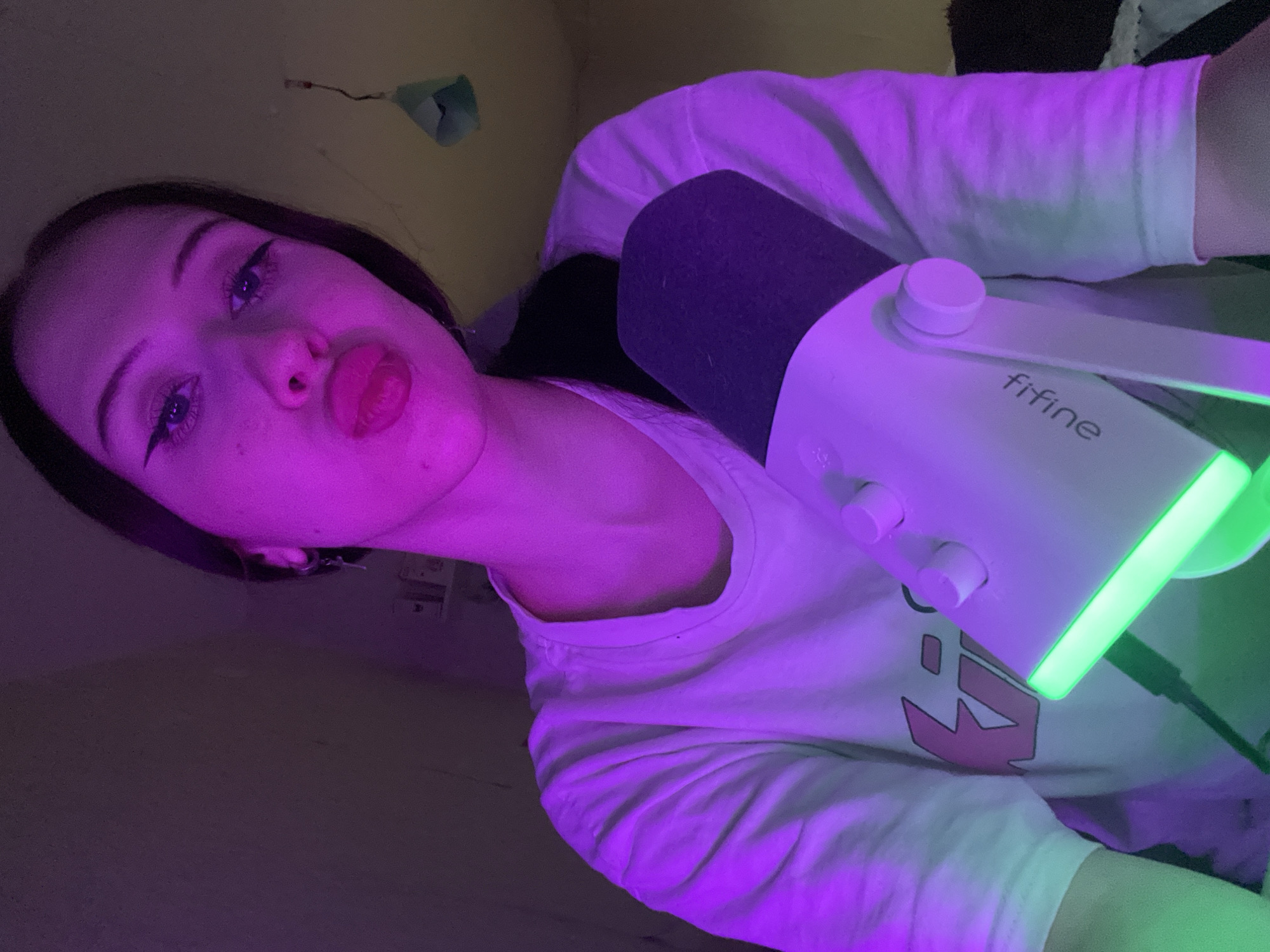 This is a medium-shot, indoor, selfie-style photograph of a young woman. She is wearing a white t-shirt with a pink logo on the chest and has her lips pursed. She is positioned in front of a white microphone with a black grille and a green light at its base. The background is dimly lit and appears to be a wall. The lighting in the image is predominantly purple, with some green accents. The exact location within Votkinsk, Russia, cannot be determined from the provided image.