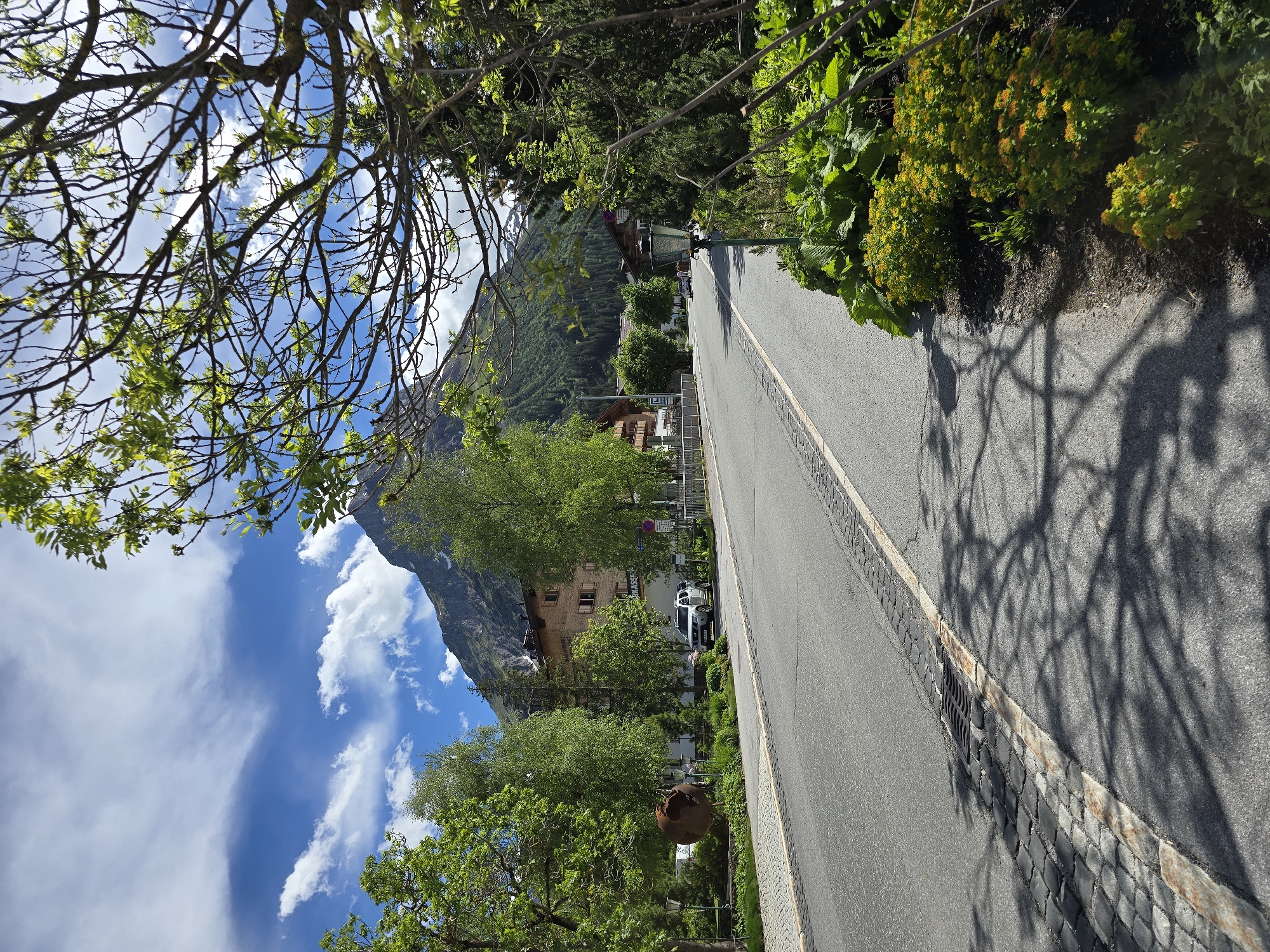 An eye-level, medium shot captures a sunlit scene on a road in Lech, Austria. The road stretches into the distance, bordered by a cobblestone path and lush greenery. A white SUV is parked on the right side of the road, near a building with an ornate facade. The background is dominated by a majestic mountain, its peak partially obscured by clouds, under a vibrant blue sky. Lush trees and foliage frame the scene, casting dappled shadows on the road.